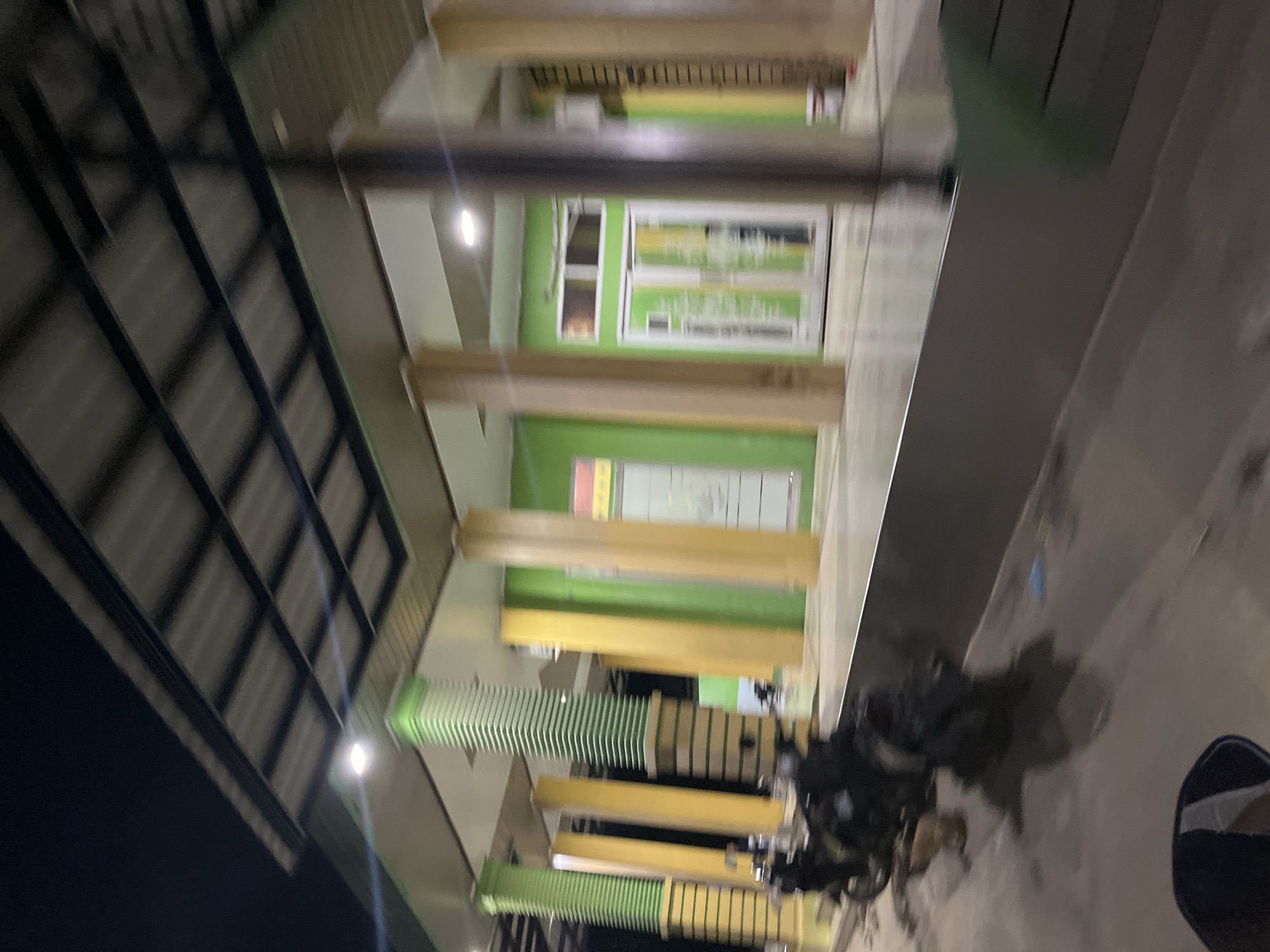 The image depicts a nighttime scene outside a multi-story building in Menanggini, Indonesia. The building features a modern design with a dark, angled roof structure supported by beams. The walls appear to be painted in vibrant green and yellow hues, interspersed with white window frames. Some windows display what appear to be posters or advertisements, though the text is not legible.
In the foreground, a cluster of motorcycles are parked on a paved surface, suggesting a common area or parking space. The lighting indicates it's nighttime, with some artificial lights visible on the building and a bright, lens-flare-like light source in the upper right quadrant. The overall impression is one of a commercial or public space, possibly an office building, community center, or retail establishment, active at night. The environment appears relatively clean and well-maintained.
