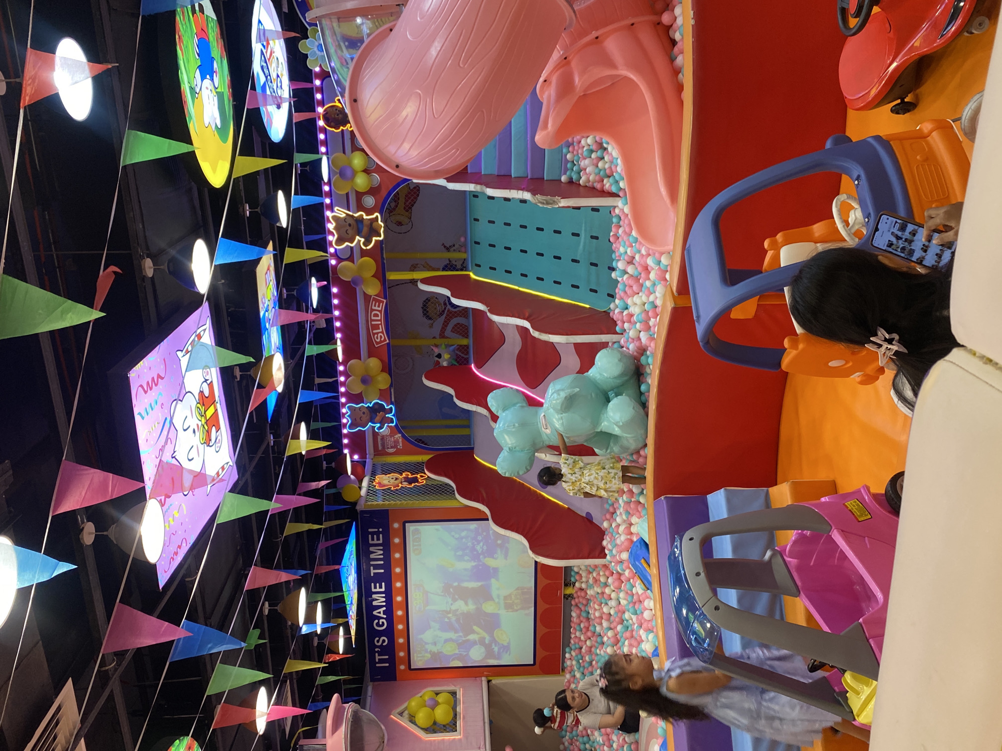 This vibrant image captures a bustling indoor children's play area, likely located within an entertainment center in Davao City, Philippines. The scene is brightly lit and adorned with numerous colorful triangular pennant banners in shades of pink, green, blue, yellow, and red, hanging from the ceiling. Illuminated cartoon character cutouts and clusters of yellow and purple balloons add to the festive atmosphere.
At the heart of the play area is a multi-level soft play structure featuring prominent slides, including a large pink slide with a wood-grain texture and a reddish-orange slide. These structures connect to a sprawling ball pit filled with countless small pink, white, blue, and light green balls.
Several children are actively engaged in play. One child, wearing a yellow and white patterned dress, stands near a large, light blue inflatable animal in the ball pit. Another child in a light blue top is seen near a pink toy car. A child with dark hair is seated in an orange and blue toy car, focused on a smartphone. An adult, possibly a parent, is also visible sitting within the ball pit, seemingly supervising or interacting with a child.
Other notable objects include various toy cars in red, orange-and-blue, and pink, and a large screen displaying animated content. Visible text on signs and decorations includes "SLIDE" on a vertical indicator, "IT'S GAME TIME!" framing the large screen, "PARADISE" on a circular overhead banner, and "HELLO" on a purple banner. The overall impression is one of a lively and engaging environment designed for children's recreation.