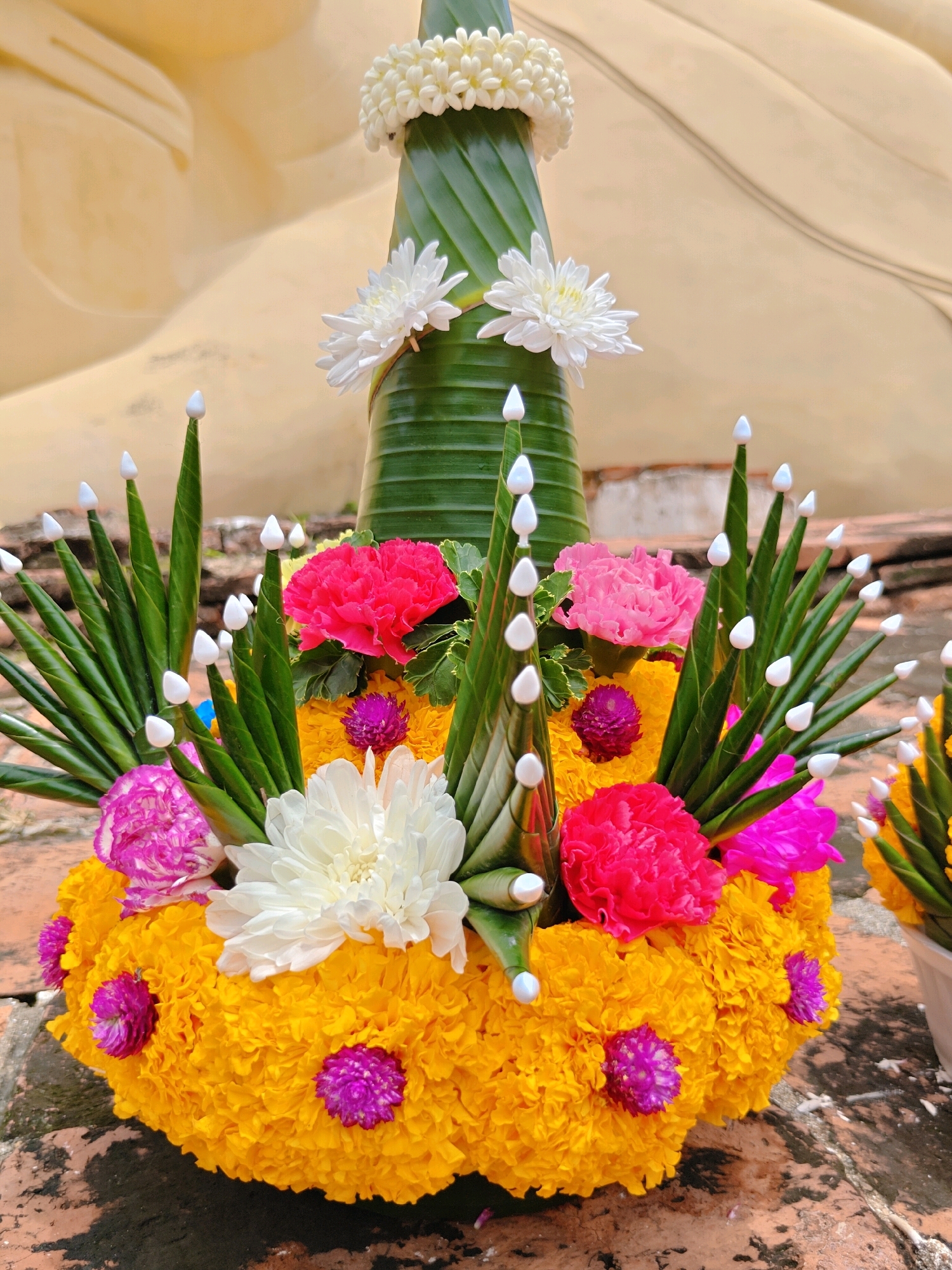 A close-up, eye-level shot captures a vibrantly decorated floral arrangement placed on an outdoor brick surface. The arrangement features a tall, conical structure wrapped in green banana leaves, topped with a white flower garland. Surrounding the base are layers of bright yellow marigolds, interspersed with pink and white chrysanthemums, and small purple globe amaranth flowers. Green banana leaf structures, adorned with white pointed tips, stand upright around the bouquet. In the soft-focus background, a large, pale yellow, curved surface suggests the presence of a reclining Buddha statue, a common feature in temples within Phra Nakhon Si Ayutthaya City Municipality, Thailand. The lighting is bright, with natural light illuminating the scene.