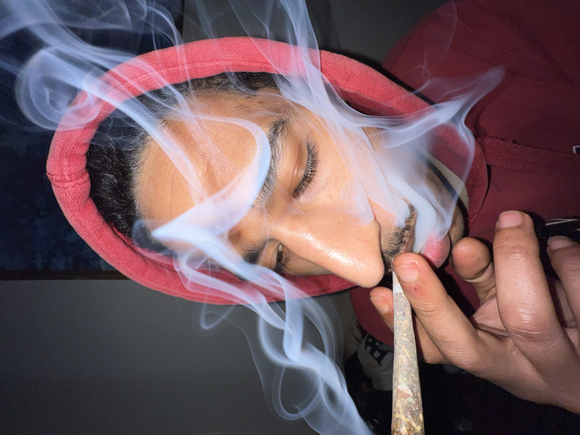 A close-up image captures a male individual in an indoor setting, located in Tanger, Morocco. The subject exhibits a medium skin tone, dark hair, and a mustache. He is wearing a dark red hooded sweatshirt, with the hood up, framing his head. The individual is actively smoking a hand-rolled cigarette, held between the index finger and thumb of his right hand and positioned at his mouth. Significant plumes of white smoke are visible, emanating from the subject's mouth and swirling around his face. His eyes are closed or heavily squinted. The hand-rolled cigarette appears to be constructed from a light-colored rolling paper with visible plant material inside. The background is dark and indistinct, with a partial view of a blurred, dark blue element to the upper right and a light-colored object with dark lettering visible in the lower-right quadrant, consistent with an interior environment.