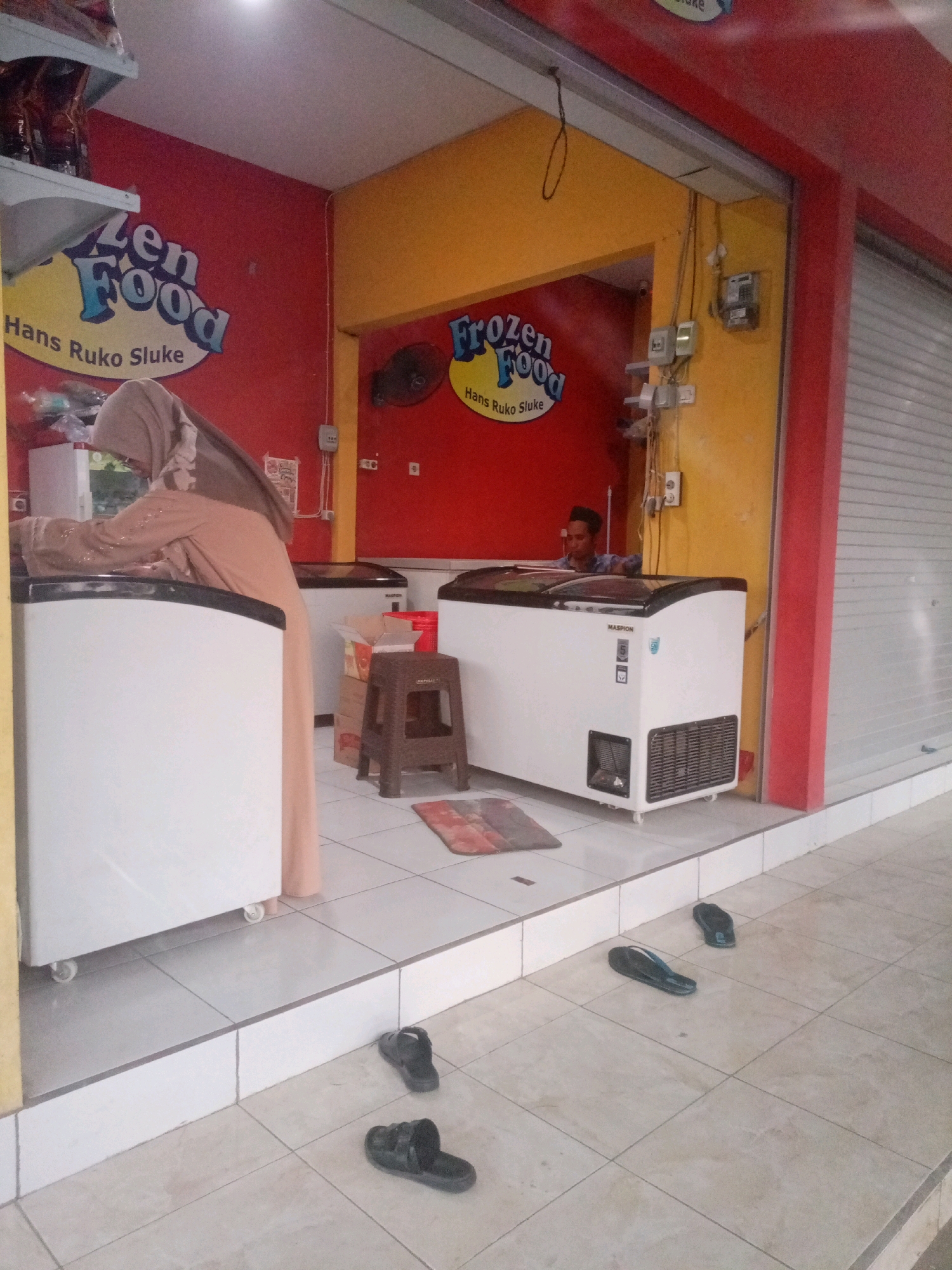The image depicts a storefront in Sluke, Indonesia, identified by a sign reading "Frozen Food Hans Ruko Sluke." The storefront is painted in vibrant red and yellow. Inside, two large chest freezers are prominent. A person wearing a beige hijab and a matching long-sleeved dress is standing by the left freezer, appearing to be tending to it. Another person, a young man with short dark hair wearing a blue plaid shirt, is visible behind the right freezer, looking towards the camera. Several pairs of black sandals are scattered on the tiled floor outside the storefront, suggesting a casual environment. Various electrical outlets and meters are mounted on the yellow wall. The scene suggests a retail establishment selling frozen goods in a local Indonesian setting.