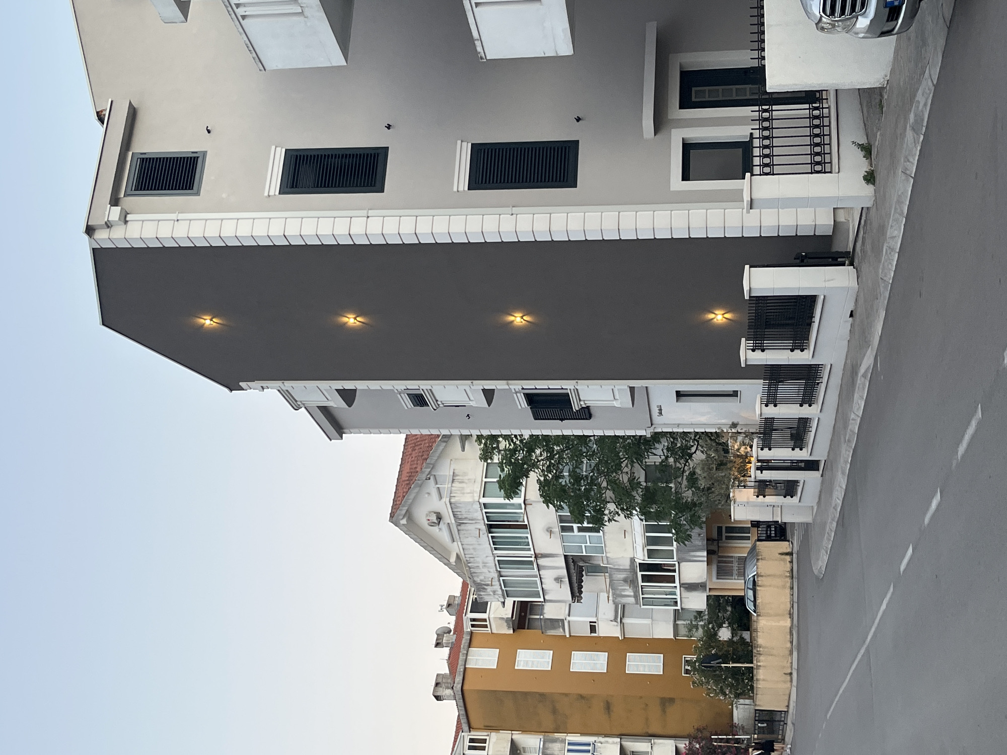 The image depicts a street scene in Budva, Montenegro, with several multi-story residential buildings lining the road. The architecture varies, with one building featuring an orange facade and white shutters, another with a cream-colored exterior and numerous balconies, and a modern grey building with minimalist design elements. The grey building has distinct vertical white trim and four upward-facing exterior lights arranged in a column on its dark grey section. A black metal fence with white pillars separates the property from the sidewalk and street. A silver SUV is partially visible on the right side of the frame. The sky is a pale blue, suggesting it is either early morning or late evening. The road has white lane markings.