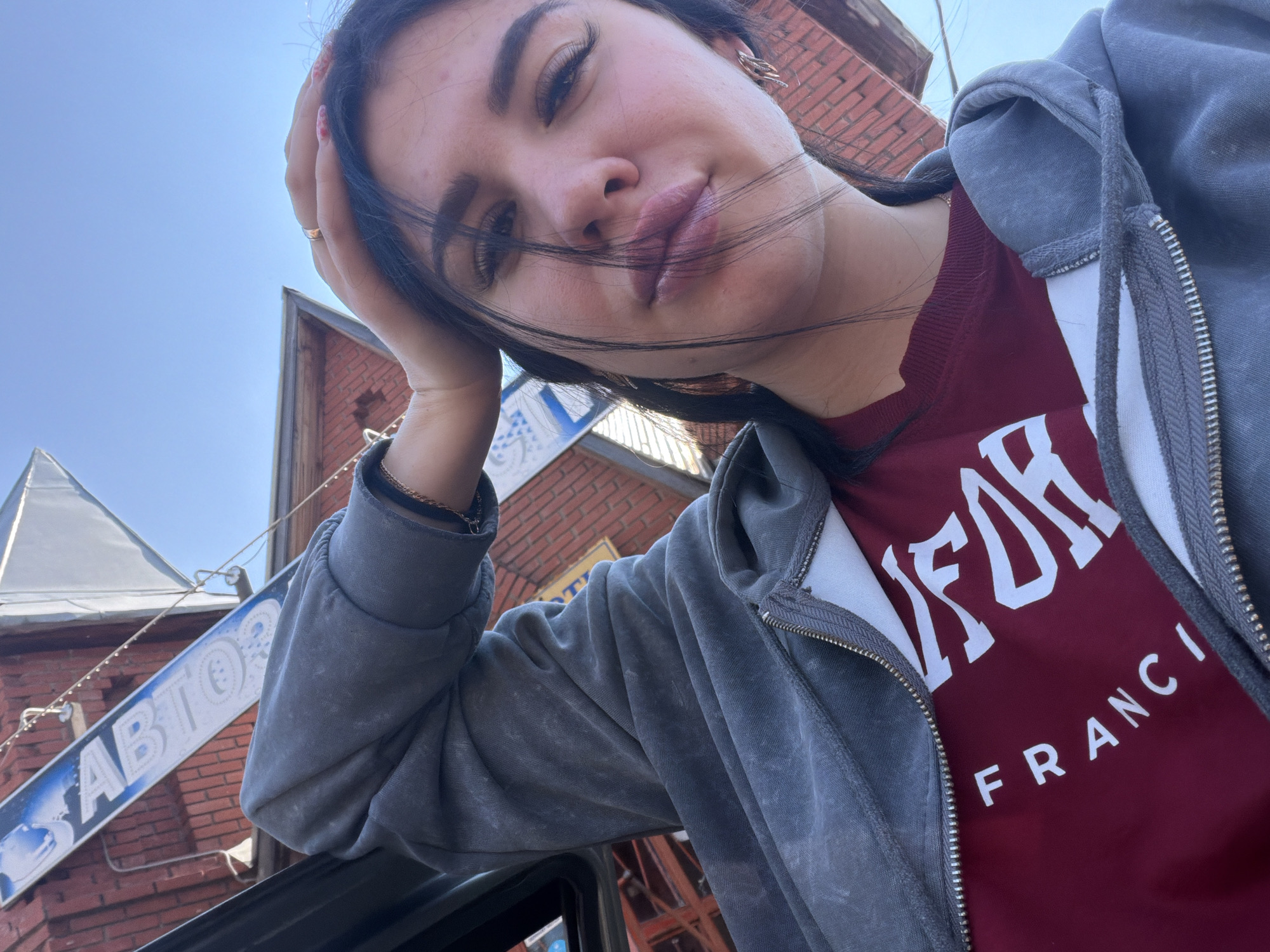 A young woman with dark hair and full lips, wearing a gray zip-up hoodie over a maroon shirt with white lettering, is captured in a close-up, tilted selfie. Her right hand is raised, resting on her head, and she wears a gold ring on her right ring finger and gold earrings. The background shows a brick building and a clear blue sky. A sign with blue lettering is partially visible on the left. The context of this image is not within the city of Фоки, Russia; it is an outdoor scene.