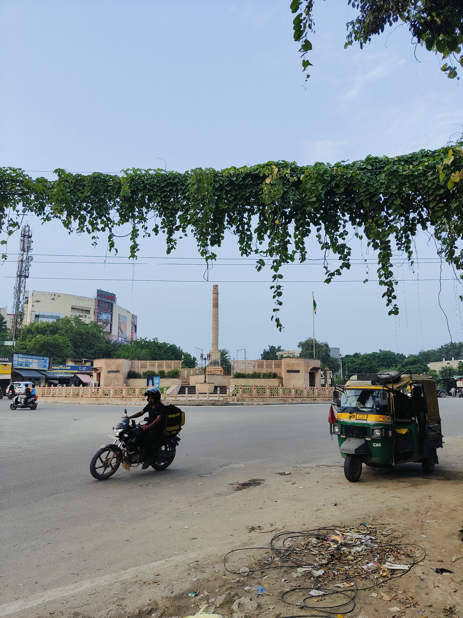 The image captures a street scene in Gurgaon, India. In the foreground, a man is riding a motorcycle with a yellow delivery box attached to the rear. To the right, a green and yellow auto-rickshaw is parked. The street is paved, and a pile of debris, including wires and trash, is visible on the side. In the background, a tall, slender monument or pillar stands prominently, with a building featuring billboards behind it. A flagpole with a green flag is also visible. Lush green vines hang from an overhead structure, partially obscuring the sky. The overall atmosphere suggests an urban environment with daily life and traffic.