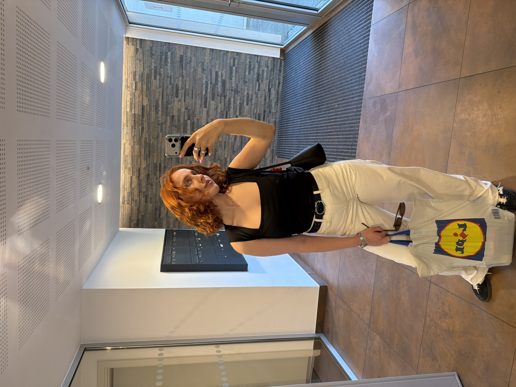 A young woman with short, red, curly hair stands in a hallway and takes a mirror selfie. She is wearing a black tank top, a black belt, and white pants. She has a black purse slung over her shoulder and is holding a white shopping bag with the Lidl logo on it, along with a pair of sunglasses. The hallway has tiled floors, a wall with mailboxes, and large glass doors at the end. The ceiling is made of acoustic tiles. The image was taken in Strasbourg, France, likely in a residential building or an office space.