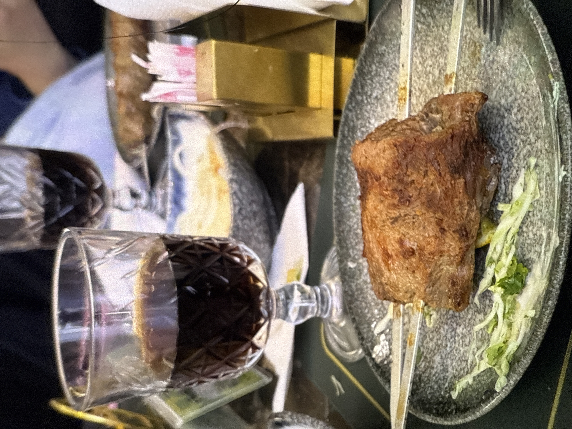 The image shows a close-up of a dining setting with a focus on food and drink. In the foreground, a patterned glass with dark red liquid, likely wine, is prominently displayed. To the right, a piece of grilled meat, possibly steak or lamb, is skewered and served on a textured, dark plate. A small portion of shredded lettuce accompanies the meat.
In the background, elements suggest a meal or gathering. A second glass, also appearing to hold red wine, is partially visible to the left. Behind the food and drinks, there are glimpses of other objects, including what appears to be a golden box or container and some pink and white items, possibly napkins or decor. The overall lighting suggests an indoor setting, possibly a restaurant or a home dining area. The image has a slightly grainy quality, common in low-light photography or older digital images. No people are directly visible, but the presence of tableware and food implies human activity related to dining. The scene appears to be captured during a mealtime, with the food ready to be consumed.