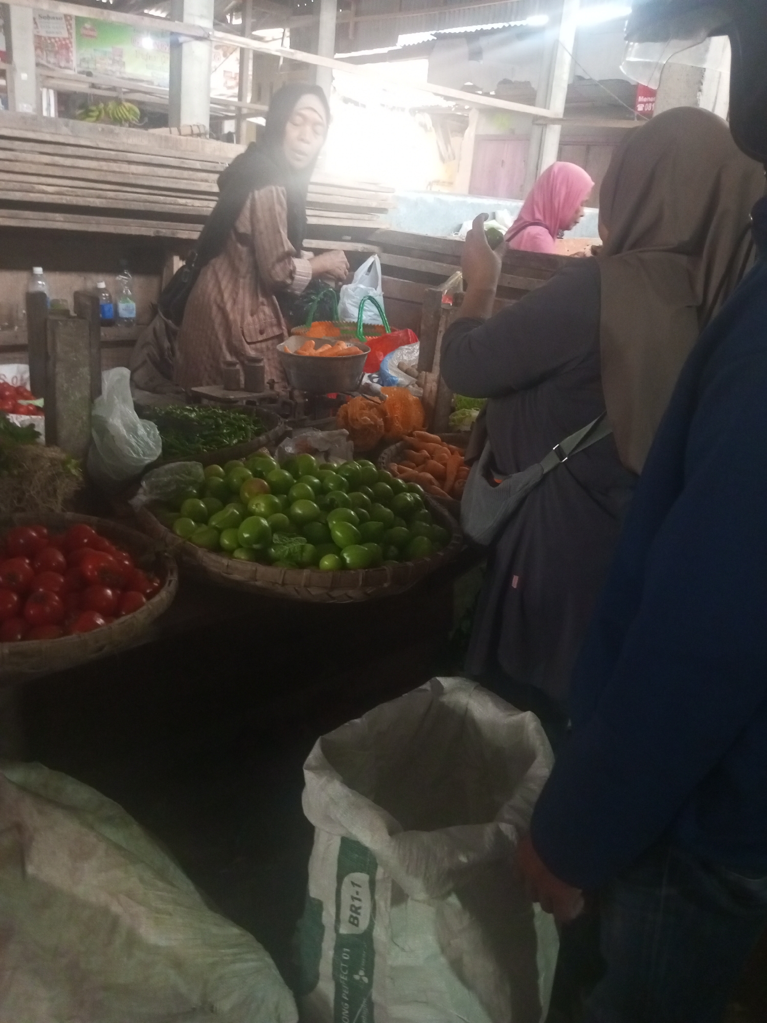 The image captures a bustling scene at a market in Kalibeber, Indonesia. In the foreground, a woman in a dark hijab is sorting green tomatoes from a woven basket. To her left, another basket overflows with ripe red tomatoes. Behind her, a woman in a pink hijab and a woman in a brown hijab are engaged in activity, with the latter appearing to be handling produce. A variety of vegetables, including carrots, are visible on the market stalls. The market appears to be an open-air or covered structure, with wooden planks and corrugated metal roofing visible in the background. The overall impression is one of vibrant market activity.
