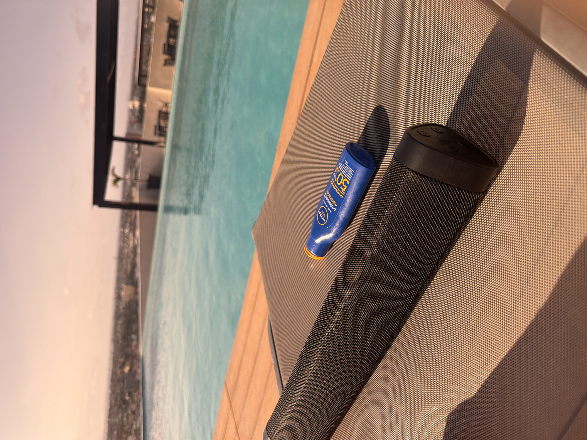 The image depicts a scene at a rooftop swimming pool overlooking the city of Tampico, Mexico. The camera's perspective is from an elevated position, looking down at a portion of a woven sun lounger with a bottle of sunscreen and a black wireless speaker on it. The sunscreen bottle is blue and yellow with "SPF 50" visible on the label. The wireless speaker has a cylindrical body and a black circular top, with several buttons visible. The pool water is a clear turquoise color, and the city skyline is partially visible in the background, framed by a black structural element. The image seems to be taken during the daytime, with natural light casting shadows.