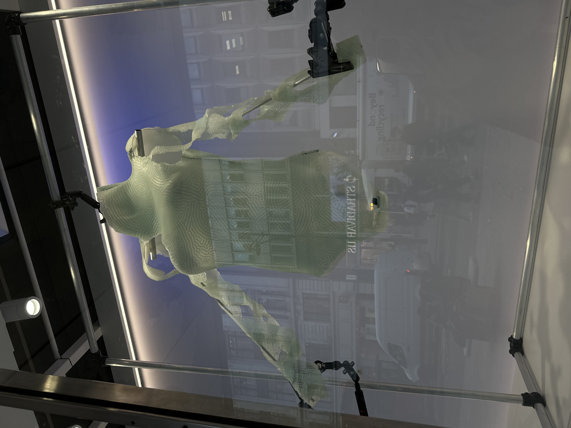 A three-dimensional, semi-transparent, dress-like sculpture with a maze-like pattern is displayed in a clear, possibly glass or plastic, case. The structure supporting the sculpture is composed of metal rods and joints. The backdrop behind the sculpture is a blurred urban scene with visible buildings and street activity, suggesting a location in a city. The reflection on the display case reveals buildings characteristic of London, United Kingdom. The sculpture is held in place by mechanical arms or fixtures at the top and bottom, appearing to suspend the artwork. The lighting is bright, highlighting the details of the sculpture.