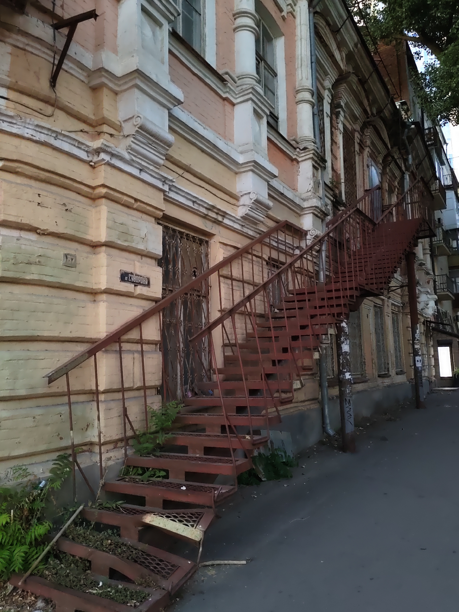 The image captures an exterior view of a historic building in Rostov-on-Don, Russia. A prominent rusty metal fire escape dominates the foreground, leading up to an entrance on the building's facade. The building itself appears to be an older apartment building, with a pale yellow painted brick lower level and a pink upper level. Architectural details, such as decorative moldings and columns, are visible. A street sign with Cyrillic lettering, reading "Суворова" (Suvorova), is affixed to the wall near the fire escape entrance, indicating the location is likely on Suvorova Street. Vegetation, including ferns and other plants, has grown around the base of the fire escape and on some of the lower steps, suggesting a degree of neglect. The surrounding environment appears to be a city street with asphalt paving in the foreground and a glimpse of trees and other buildings in the background. The overall impression is one of aged architecture and urban decay.