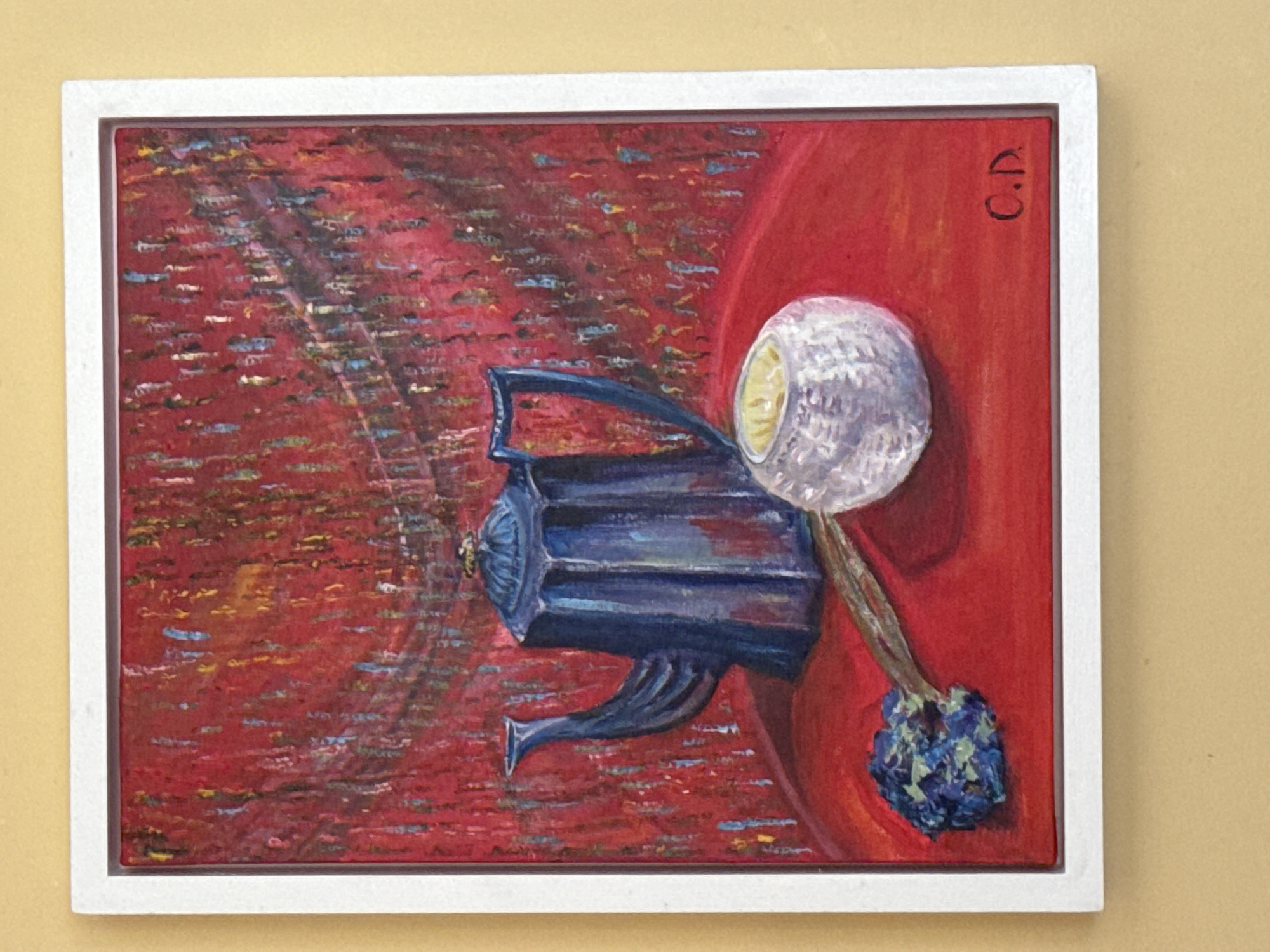 The image shows a framed painting of a still life. The painting depicts a blue teapot, a white textured object that appears to be a candle holder with a lit candle, and a small bunch of blue flowers on a red surface. The background of the painting is a textured red with streaks of other colors. The artist's signature, "C.D.", is visible in the lower right corner of the painting. The painting is framed with a white border and is hanging on a yellow wall. The image does not provide any information about its location in the city of Waterford, Ireland.