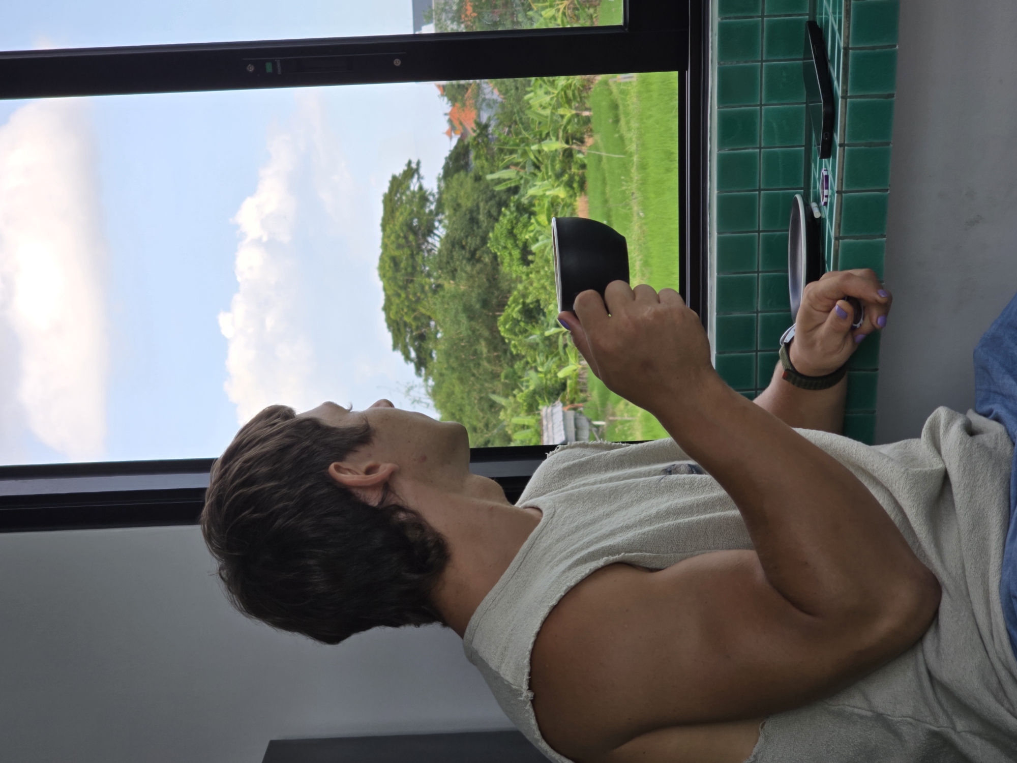 A person sits in a room in North Kuta, Indonesia, holding a black mug and looking out of a window. The window frames a view of green trees and rice paddies under a blue sky with white clouds. The person has short brown hair, a fair complexion, and is wearing a sleeveless white shirt. They have a watch on their left wrist and purple nail polish. The wall next to the window is covered in green square tiles, and a phone is affixed to the tile wall. The room is lit by natural light coming through the window.