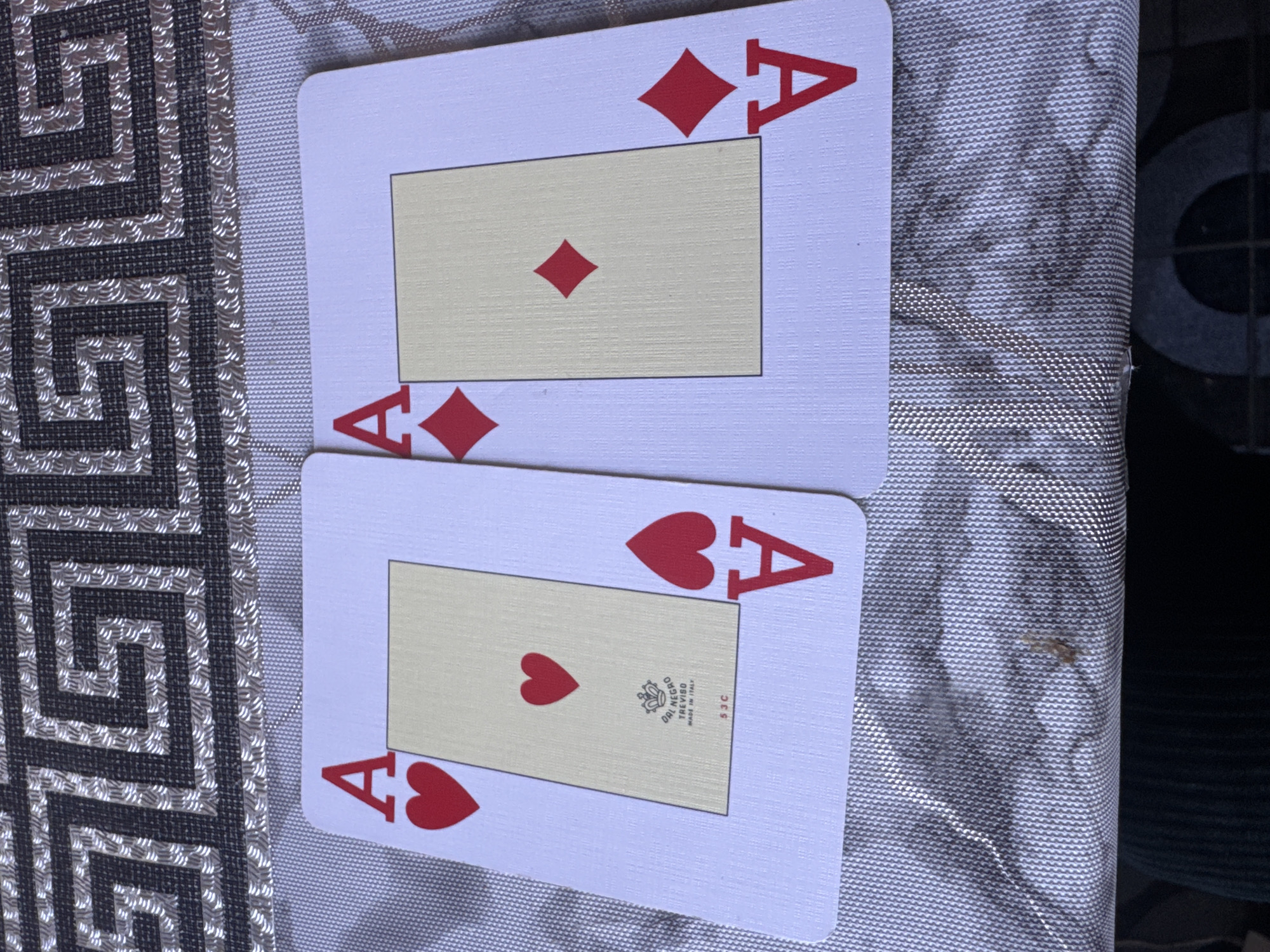 Two Aces are laid out on a table with a patterned tablecloth. The Ace on the left is the Ace of Hearts, and the Ace on the right is the Ace of Diamonds. The background tablecloth has a geometric pattern. There are no people or actions depicted in the image. The scene appears to be indoors. The location in Zaokskiy, Russia cannot be determined from the image.