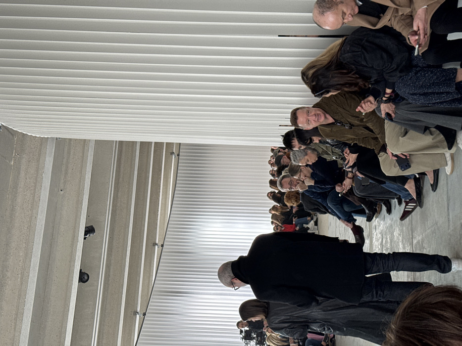 The image displays an indoor event, likely a fashion show or presentation, set within a contemporary, minimalist space in Paris, France, possibly an exhibition hall such as the Palais de Tokyo or a similar adapted venue. The architectural elements include a high, exposed concrete ceiling with visible structural beams and black track lighting fixtures. The walls are constructed from tall, curved, white vertical fluted panels that extend into the background, creating a sleek, structured environment.
Approximately 20-30 adults are clearly visible, primarily seated in multiple rows on the right side of the frame, facing an unseen central event to the left. In the immediate foreground, a man wearing a dark coat and glasses stands with his back to the camera, beside a woman in a long black coat. The seated attendees wear a range of attire. Many individuals are interacting with smartphones; some are viewing content, while others hold their devices ready. A man in a brown jacket and tan trousers is prominent in the front row of the seated audience, looking to the left. In the far background on the left, a person operates a professional video camera mounted on a tripod. The floor is a light, unadorned surface, consistent with concrete.