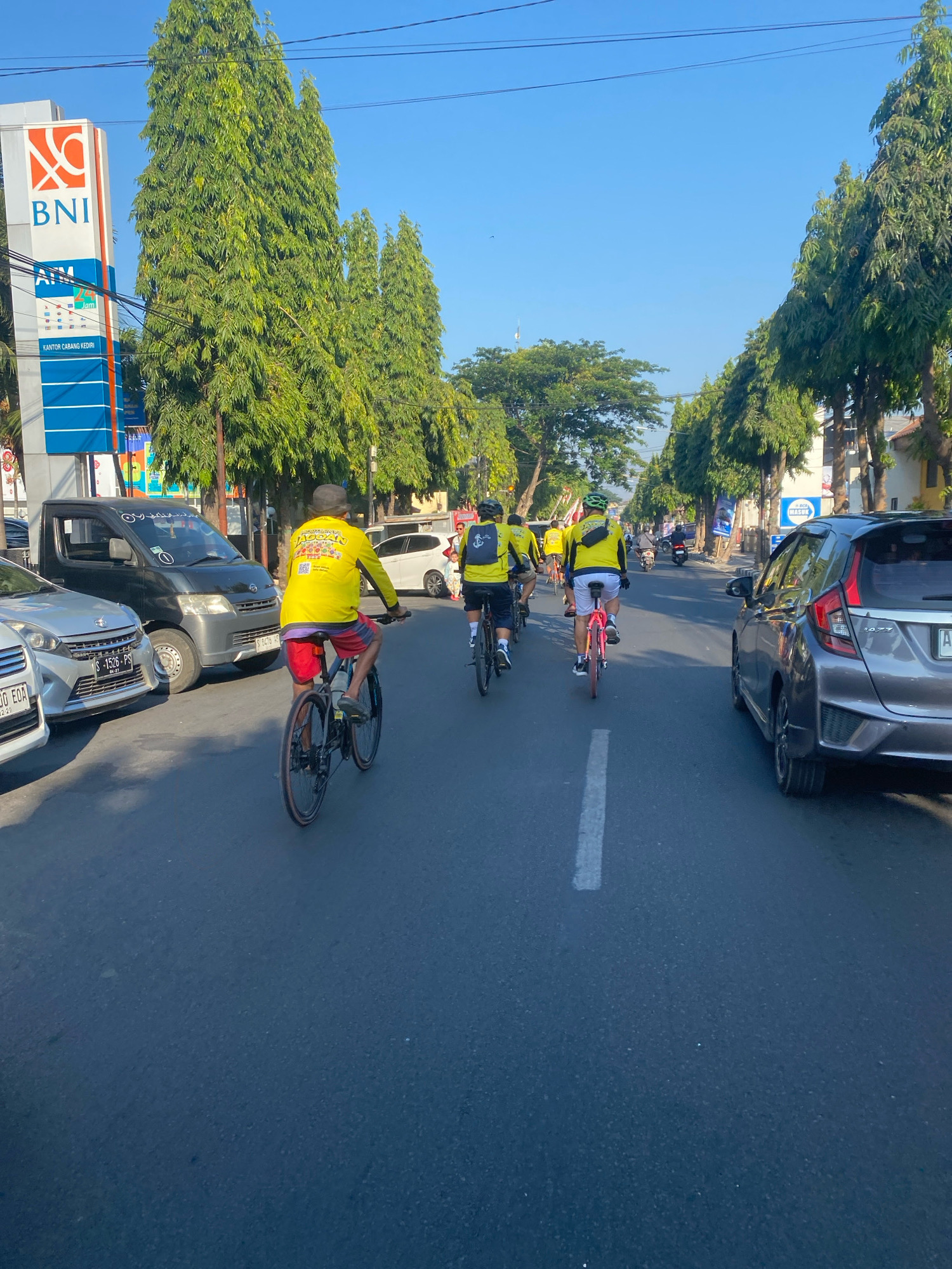 A group of cyclists wearing bright yellow shirts are riding on a paved road in Kediri City, Indonesia. The cyclists are in the middle of the road, with cars and a black truck to their left. Tall, slender trees line the side of the road, and a bright blue sky is visible above. A BNI bank sign is prominent on the left side of the image. The cyclists appear to be part of an organized event, given their matching attire and the presence of other cyclists further down the road. The scene suggests a sunny day with good visibility, typical of a morning in Kediri.