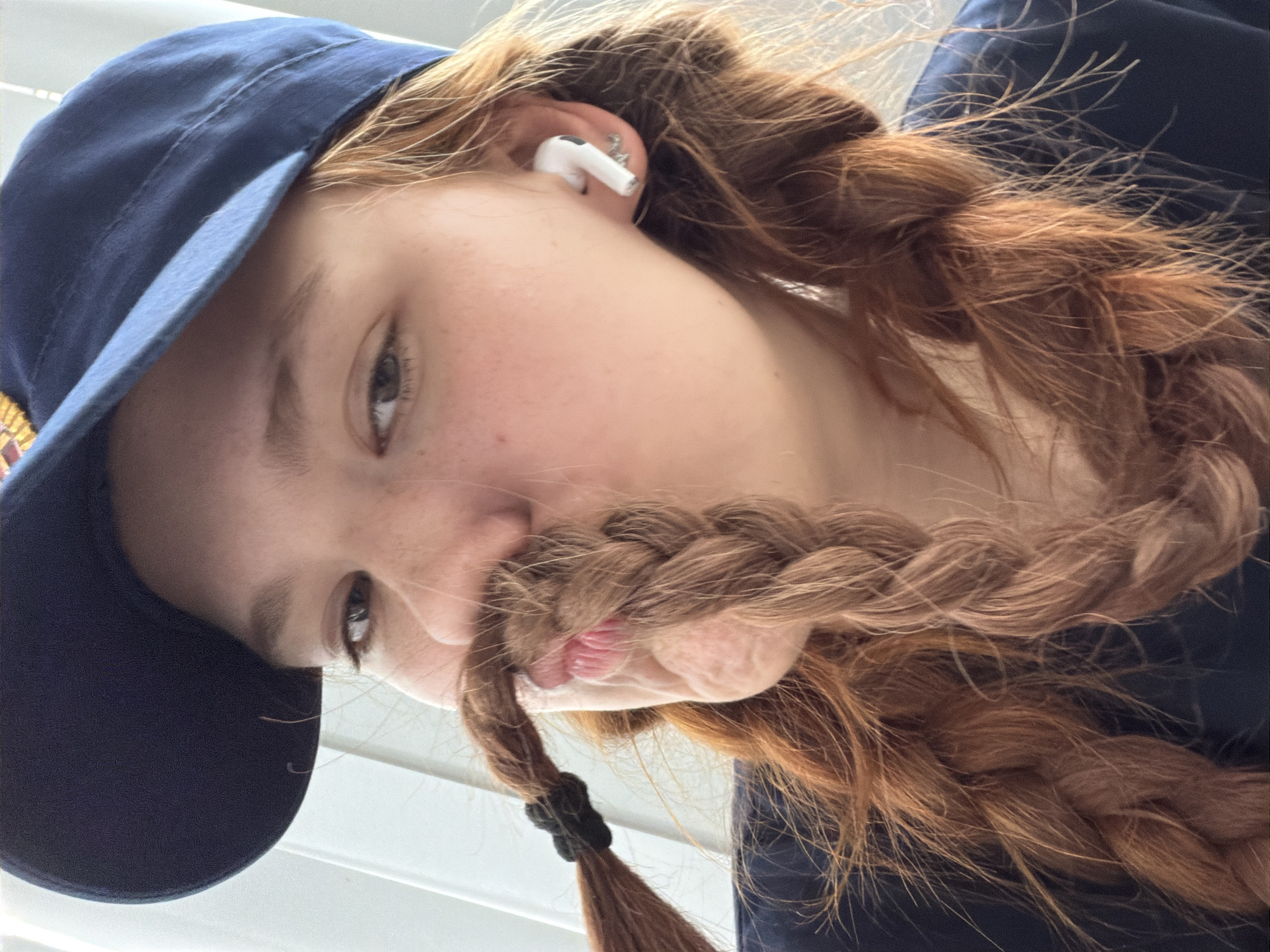 The image shows a close-up, low-angle, sideways shot of a young person with red hair wearing a navy blue baseball cap. The person has a braid of hair draped across their face, obscuring their mouth and nose, and appearing like a mustache. A white earbud is visible in their left ear. The background is out of focus, showing white horizontal blinds and a hint of blue fabric, possibly clothing. The lighting suggests it is daytime. The overall mood is playful and informal. No text is visible.