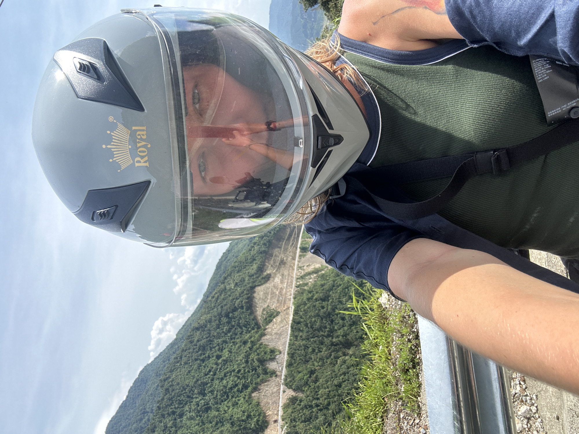 An overhead, slightly angled full-shot image, originally rotated 90 degrees clockwise, captures a person wearing a motorcycle helmet against a dramatic mountainous backdrop. The subject, likely taking a selfie, wears a grey full-face helmet with a clear visor. The brand "Royal" and a crown logo are visible in gold lettering on the side of the helmet. Through the clear visor, the person's eyes (greenish/hazel) are visible, along with reflections of their face and possibly the camera device. Wisps of light-colored hair peek from beneath the helmet.
The person is dressed in a dark green ribbed tank top, over which a dark blue/navy short-sleeved top is worn. A black strap, possibly from a backpack or the helmet's chin strap, crosses the chest. A clothing tag is visible on the green top, showing text including "MADE IN CHINA", "100% LINEN", "S Bust 77-83cm", and "身高 155-160cm", along with Chinese characters and the number "801-4217200". A bare arm, resting on a silver metal guardrail at the bottom right of the frame, shows fair skin and a faint tattoo on the upper arm.
The scene depicts a sunny day with a clear blue sky and scattered white clouds. The background is dominated by lush green mountains, suggesting a natural and rugged landscape. A wide, light-colored path or road is visible cutting through the mountainside, indicating a passage through the dense forest. The setting points to a scenic viewpoint, likely in Muong Than, Vietnam, given the context. The time of day appears to be bright daylight.
