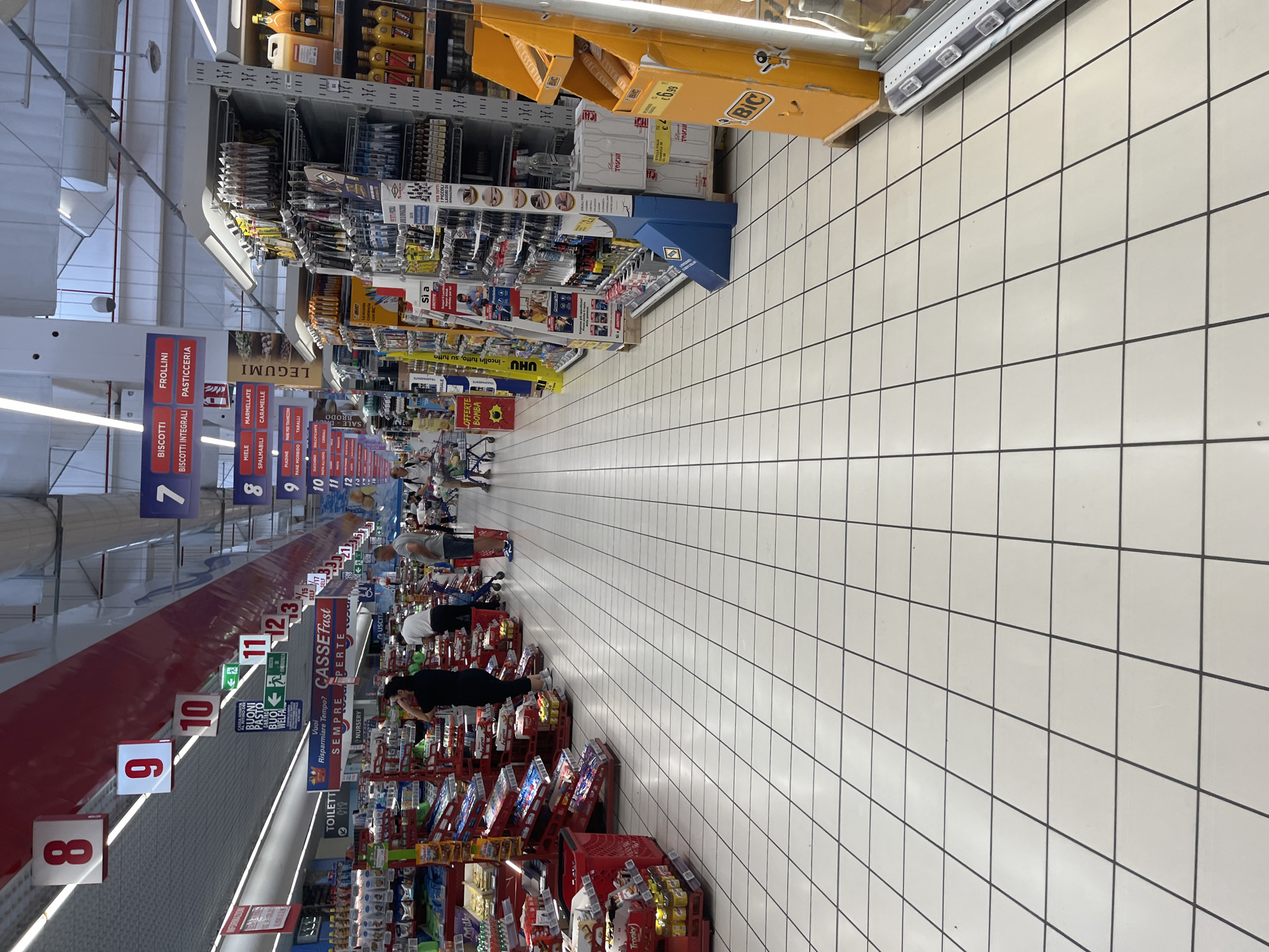 This image shows the interior of a large supermarket, likely a "Superstore" or similar hypermarket, given the scale of the aisles and the variety of goods on display. The floor is tiled with a white grid pattern. The ceiling is high, with white panels and visible light fixtures. In the distance, aisles are stocked with a wide range of products including food items, household goods, and possibly stationery or craft supplies based on the visible packaging and displays.
Several shoppers are visible throughout the store. On the left side, a person in a black t-shirt and jeans is standing, possibly looking at products. Further down, a man and woman with a shopping cart are visible. On the right side, several people are browsing shelves. A prominent orange display stand for "BIC" products is visible on the right side of the image.
Various signage is present, including directional signs, product category labels, and promotional banners. Numbers are displayed on hanging signs, such as "8", "9", "10", "11", and "12", which likely indicate aisle numbers or sections within the store. One sign with green arrows indicates an emergency exit. Other signs visible include "BISCOTTI", "FROLLINI", "PASTICCERIA", "LEGUMI", and "TOILETTI".
The overall impression is of a busy, well-stocked supermarket. Without further information or distinct landmarks, pinpointing the exact location within Castiglione delle Stiviere is not possible based solely on the image. However, the store appears to be a typical large-format retail establishment.