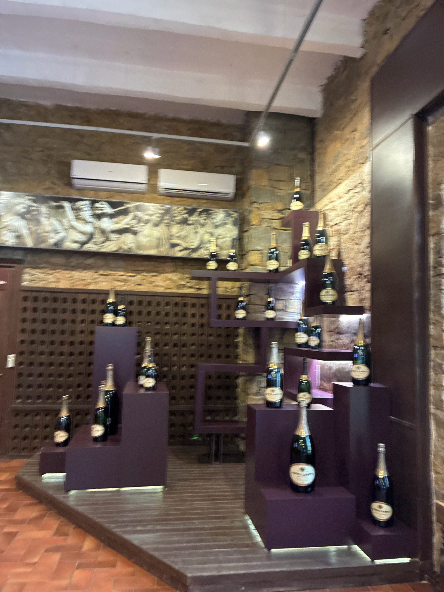The image shows a display of champagne bottles in a rustic interior. The walls are made of rough-hewn stone and rough plaster. There are two air conditioning units mounted high on the wall. A large bas-relief artwork depicting figures is mounted above a wooden lattice-patterned wall. The champagne bottles are displayed on tiered purple risers against the stone wall. The floor is a combination of dark wood planks and terracotta tiles. The scene is likely located in Abrau-Dyurso, Russia, a region known for its vineyards and champagne production. There are no people visible in the image.