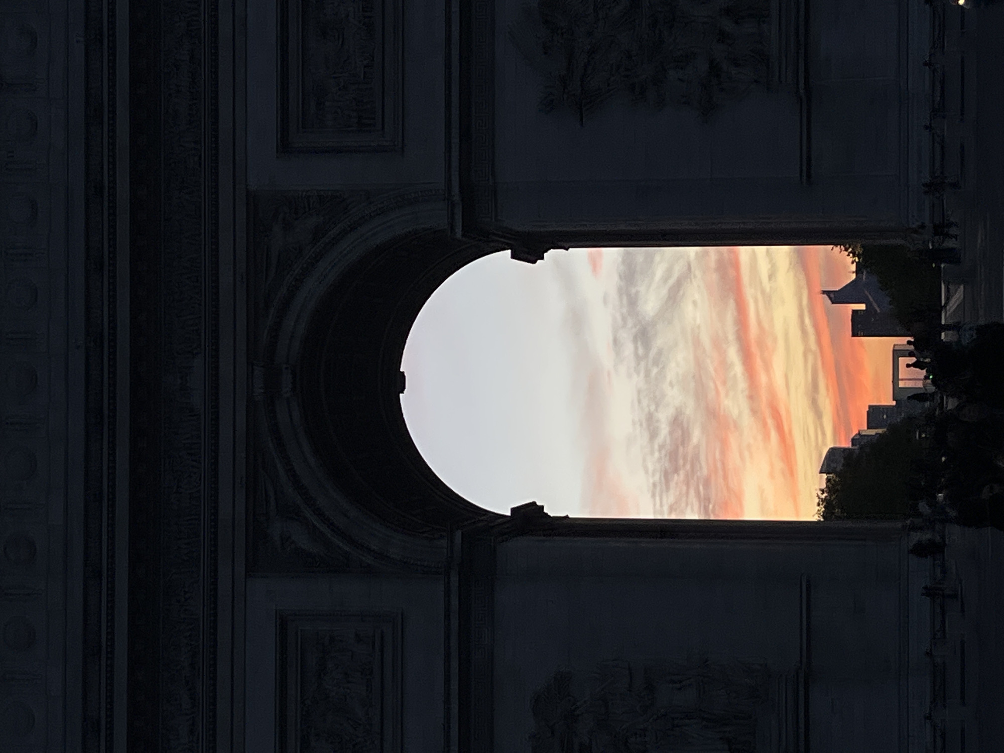 The image displays a monumental archway, characteristic of the Arc de Triomphe, framing a vibrant sunset or sunrise sky. The photograph is oriented vertically, with the arch occupying the left side of the frame, and the vista through it on the right.
The arch, presumed to be the Arc de Triomphe in Paris, France, is rendered in deep shadow, highlighting its massive dark stone structure. Intricate relief carvings and decorative patterns, depicting figures and ornamentation in a classical style, are visible across its surfaces, particularly on the recessed panels and cornices, despite the low light. The arch's imposing silhouette creates a strong contrast with the bright background.
Through the archway's large, rectangular opening, a spectacular sky is visible. On the left, the sky is a bright, luminous white or pale yellow, gradually transitioning into a rich array of orange, pink, and deeper yellow clouds on the right. These streaky clouds suggest either a dramatic sunset or sunrise.
Below the colorful horizon, on the far right, the dark silhouettes of distant city buildings and trees are outlined against the bright sky. Faint shapes of people can also be discerned on the ground level, suggesting a public space or street. No specific activities are clearly identifiable, and there is no visible text within the image. The scene conveys a sense of grandeur and natural beauty, typical of dusk or dawn in a historic city.