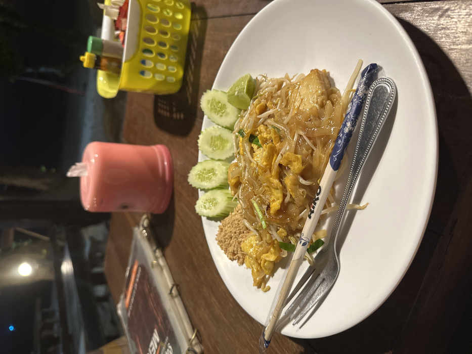 The image depicts a plate of Pad Thai served at a nighttime outdoor dining establishment in Phra Ae, Thailand. The dish, presented on a white circular plate, consists of stir-fried rice noodles with egg, bean sprouts, chopped scallions, and what appears to be chicken or tofu. It is garnished with a wedge of lime, a pile of ground peanuts, and five slices of fresh cucumber. A silver fork and a pair of white chopsticks with blue patterned tips and the brand "RIKO" printed on them rest diagonally across the plate.
The plate sits on a dark brown wooden table, which shows visible grain and texture. To the left of the plate, a menu in a clear plastic sleeve is partially visible, folded open. Behind the menu and to the right, a pink cylindrical object, possibly a decorative LED lamp or candle, emits a soft glow. Further to the right, a yellow plastic caddy with a perforated design holds small bottles of condiments (e.g., sauces, spices) and what appear to be small red fruit pieces (possibly strawberries or chillies).
The background is dimly lit, indicating an evening or nighttime setting. Blurred shapes of additional wooden tables and chairs are discernible in the distance, suggesting an open-air restaurant or a similar dining area. A faint light source is visible in the far background. No people are directly visible in the frame.