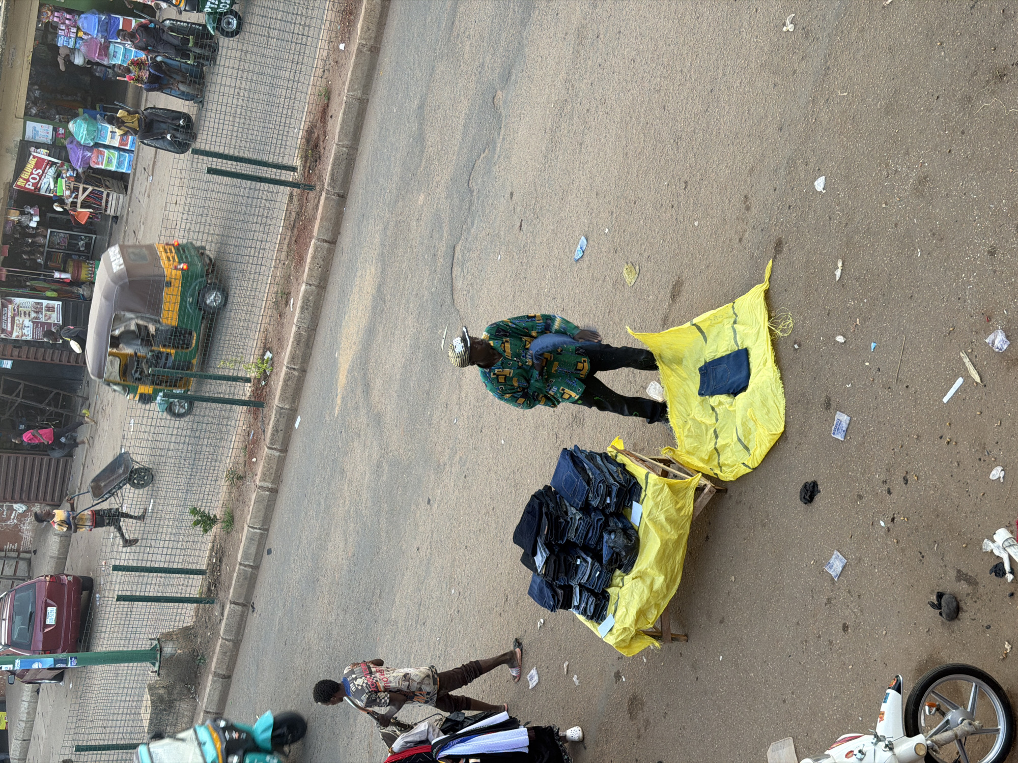 The image depicts an outdoor street scene in Bukuru, Nigeria, likely in a marketplace. A man wearing a patterned green and yellow shirt and a straw hat is standing near a pile of folded blue jeans displayed on a small wooden table covered with a yellow cloth. Another yellow cloth lies on the ground to the right, also with some folded blue jeans on it. In the background, a person walks by with a wheelbarrow, and various shops and stalls are visible, with goods displayed and people milling about. A red car is parked on the left side of the road, and several tricycles (Keke Napep) are present, one with passengers. The ground is paved, with some debris and discarded items scattered around. The scene appears to be a busy commercial area.