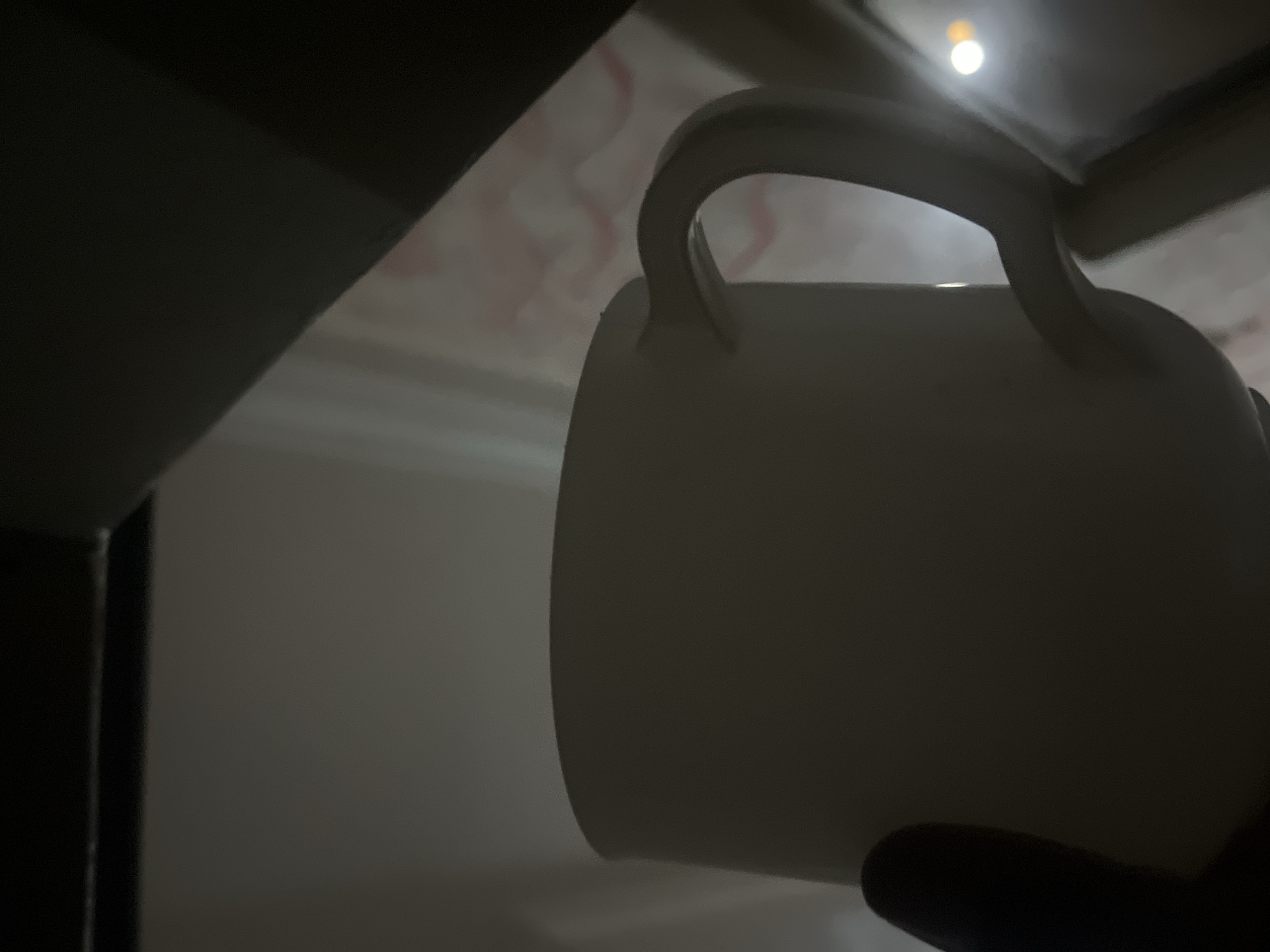 A light-colored mug with a handle is held by a dark hand in the foreground of the image. The mug is partially illuminated by ambient light, casting shadows on its surface. In the background, a blurred pattern suggests a curtain or fabric. The overall lighting is dim, with a bright circular reflection visible on a surface to the right of the mug. The scene is likely indoors, given the presence of a mug and curtain. There are no identifiable landmarks that would indicate the specific location within Jalingo, Nigeria.
