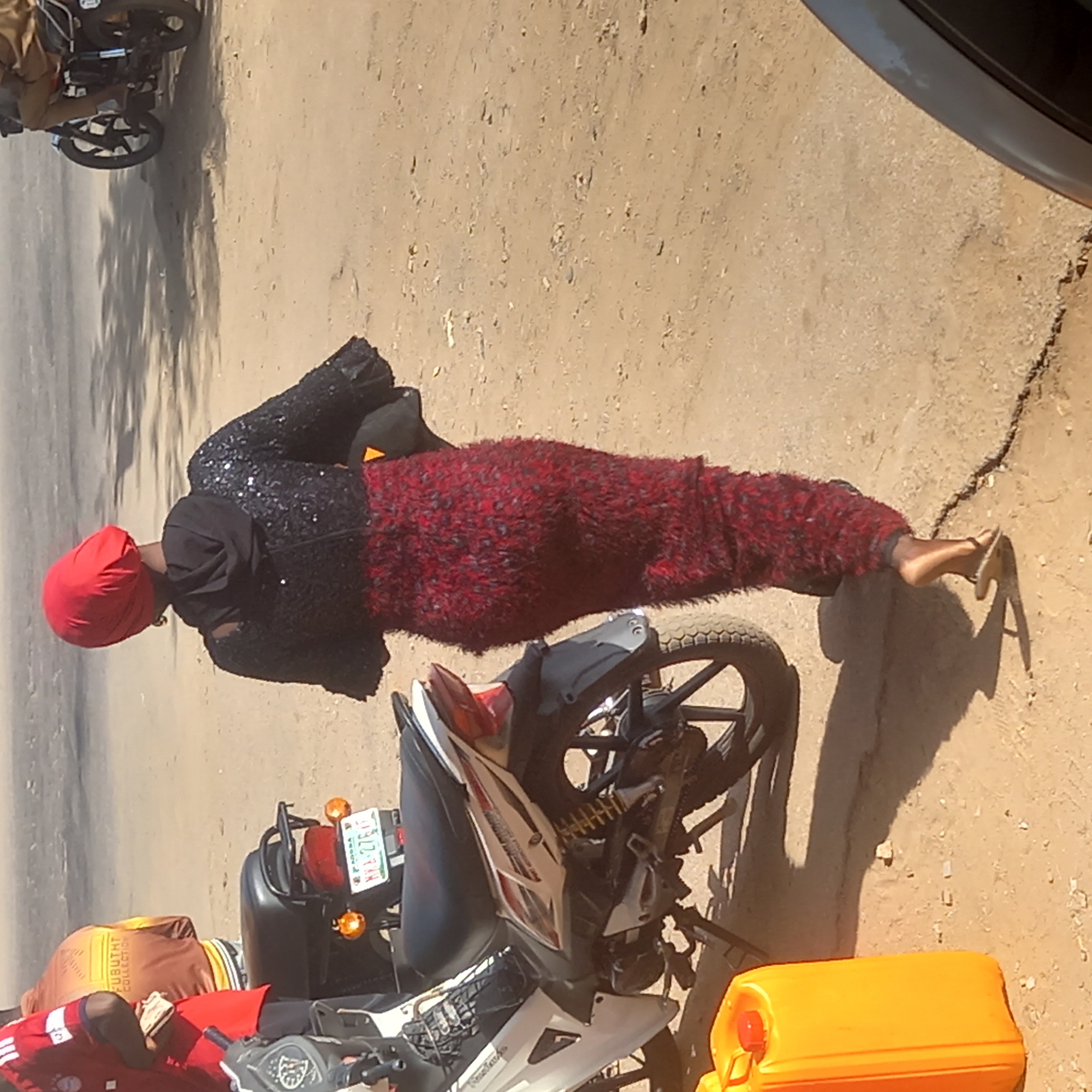 The image shows a scene on a dirt road in Sardauna, Nigeria. A woman dressed in a black sequined top, red furry pants, and a red headwrap lies prone next to a white and black motorcycle with a license plate. Another person wearing red shorts and a brown shirt lies on the motorcycle. A yellow jerrycan sits near the motorcycle. Another motorcycle is partially visible in the background.