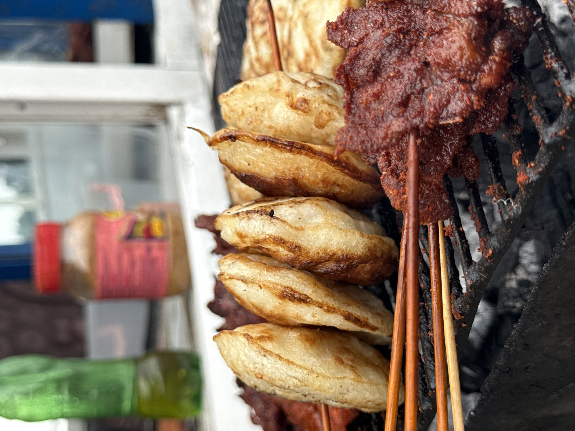 A close-up shot captures a street food vendor grilling meat and plantains on a charcoal grill in Ado, Nigeria. In the foreground, skewers of reddish, marinated meat, possibly beef or goat, are being cooked. Adjacent to the meat are several pieces of golden-brown fried plantain, also on skewers, with some appearing slightly charred. The grill grate is dark and appears to have remnants of food particles. In the blurred background, a green bottle of beverage and a jar with a red lid, possibly containing peanut butter or sauce, are visible. The scene suggests an outdoor food stall in a bustling urban environment.