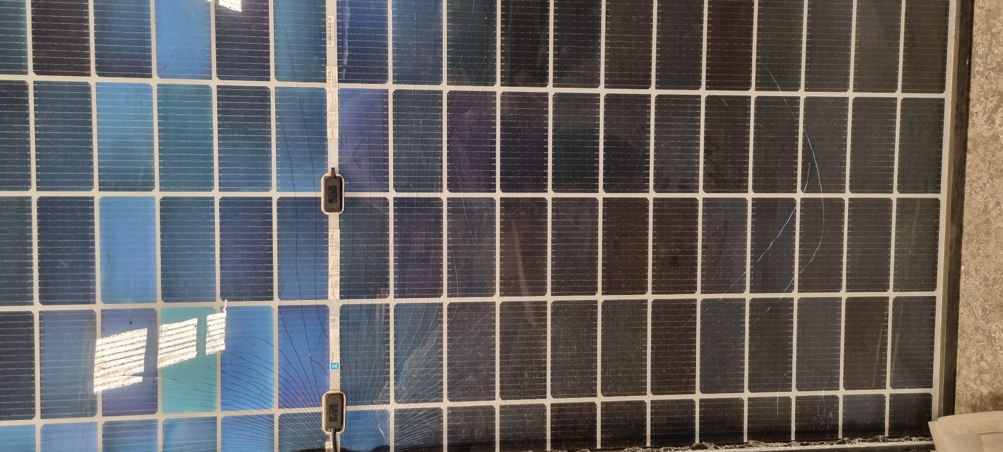 The image shows a close-up of a damaged solar panel, likely in an urban environment. The panel consists of multiple rectangular photovoltaic cells arranged in a grid pattern. The left side of the panel appears in better condition, showing reflective blue cells, while the right side exhibits darker, possibly degraded cells. Several cells display visible cracks and fractures, indicating physical damage to the panel. Two small, black boxes are attached to the panel along a vertical line, appearing to be connectors or sensors. The surrounding environment is out of focus, but a textured wall is visible on the right side of the frame.