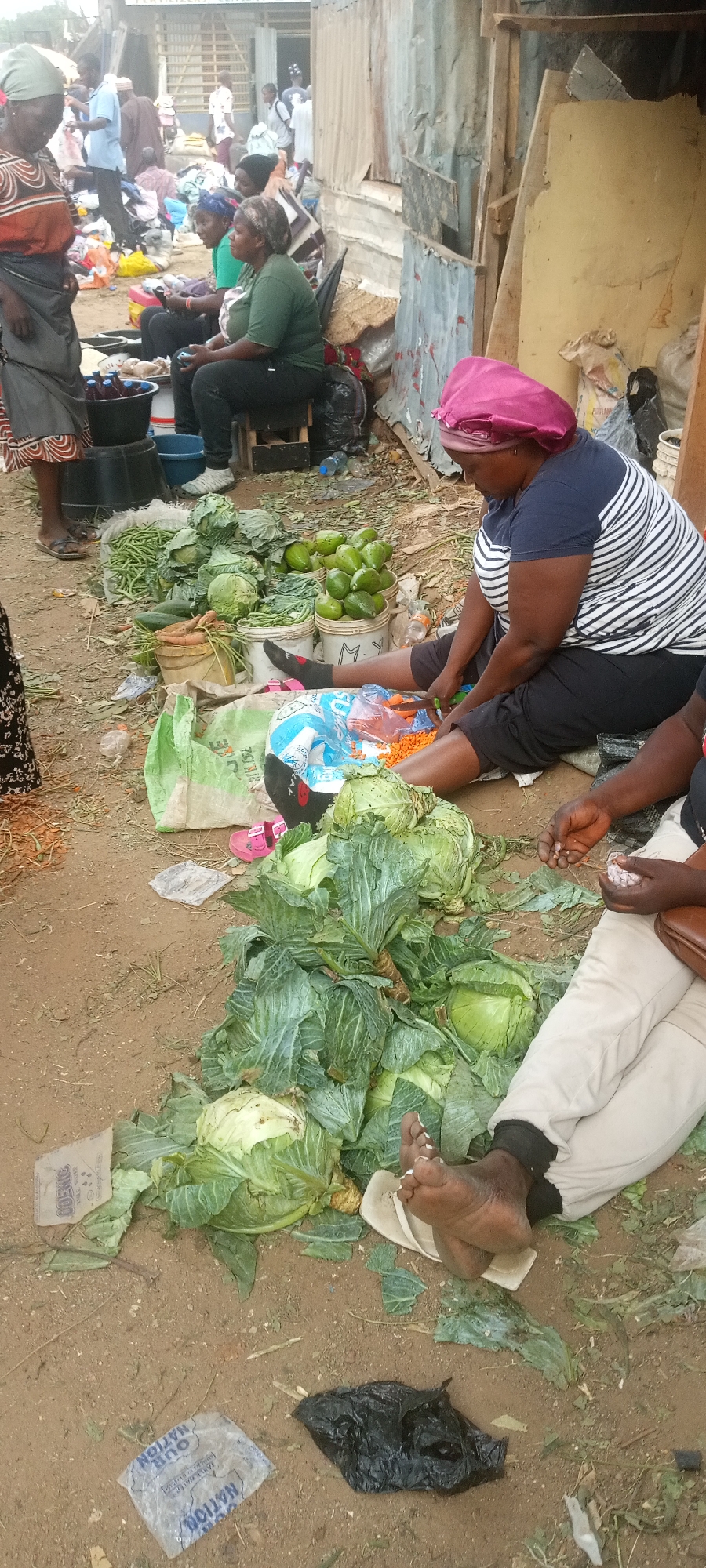 The image depicts a market scene in Gura Top, Nigeria. Several people are visible, including women selling produce on the ground. One woman is sitting and cutting carrots, while another is selling cabbages. Other produce like avocados and green beans are also displayed. The ground is dirt with scattered leaves and trash. Buildings are visible in the background.