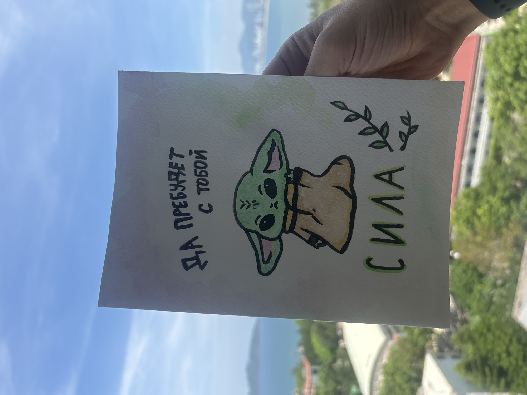 A hand holds a drawing of a green creature resembling Grogu from Star Wars. The drawing is on white paper with watercolor accents. Above the creature, text in Russian reads "ДА ПРЕБУДЕТ С ТОБОЙ" (May the Force be with you). Below the creature, the word "СИЛА" (Force) is written. The background shows a blurry view of a body of water, coastline, and buildings, suggesting an outdoor setting, possibly in a city. The sky is visible and appears blue with some clouds.