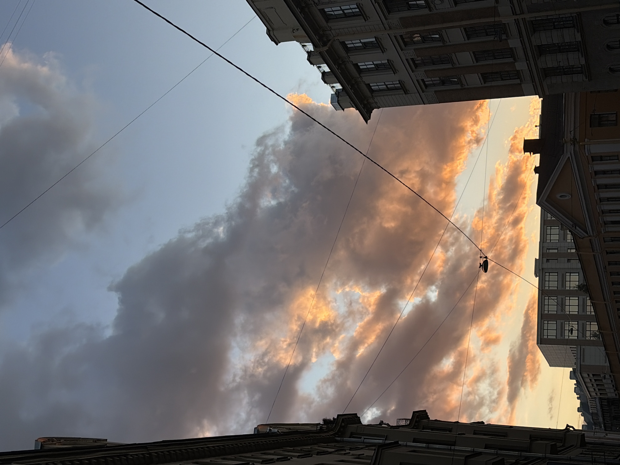 The image depicts an urban scene in Saint Petersburg, Russia, likely during sunset. The sky is filled with dramatic clouds, some of which are illuminated with warm orange and yellow hues, indicating the setting sun. The clouds are dense and layered, with patches of clear blue sky visible.
In the foreground and sides of the image, traditional Saint Petersburg architecture is visible. On the left, a dark-colored building with visible windows and architectural details runs along the edge of the frame. On the right, a lighter-colored, multi-story building with a more ornate facade and numerous windows is prominent. Further in the background, other buildings with varying architectural styles can be seen, including a modern, glass-fronted building and older structures with pitched roofs.
Several black wires, likely power or telephone lines, crisscross the sky, obscuring parts of the view and adding a sense of urban infrastructure. A dark street lamp is suspended from one of these wires in the middle ground. The overall impression is of a city street bathed in the dramatic light of a sunset, with the characteristic architectural style of Saint Petersburg providing a distinct urban backdrop. There are no people or visible actions occurring in the scene.