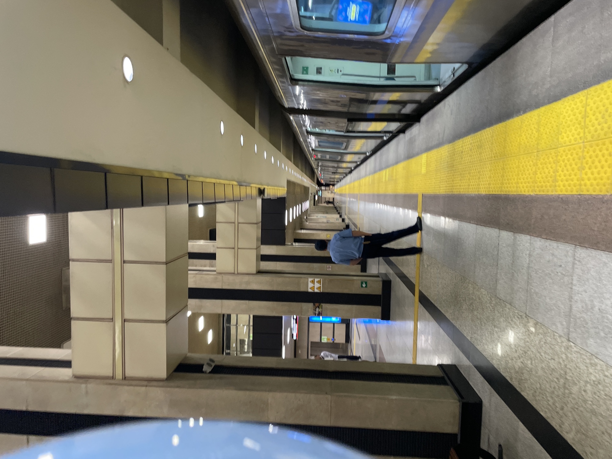 A metro station platform in Almaty, Kazakhstan, features a train with its doors open. A man in a blue shirt and dark pants stands on the platform, facing away from the camera, with his hands in his pockets. The platform is marked with a yellow tactile paving strip along the edge. The station has tall, beige, and black striped columns, a suspended ceiling with lights, and tiled flooring. In the background, other people are visible further down the platform and on an upper level. The train is modern and metallic, with visible windows and doors. The environment appears to be indoors, well-lit, and orderly.