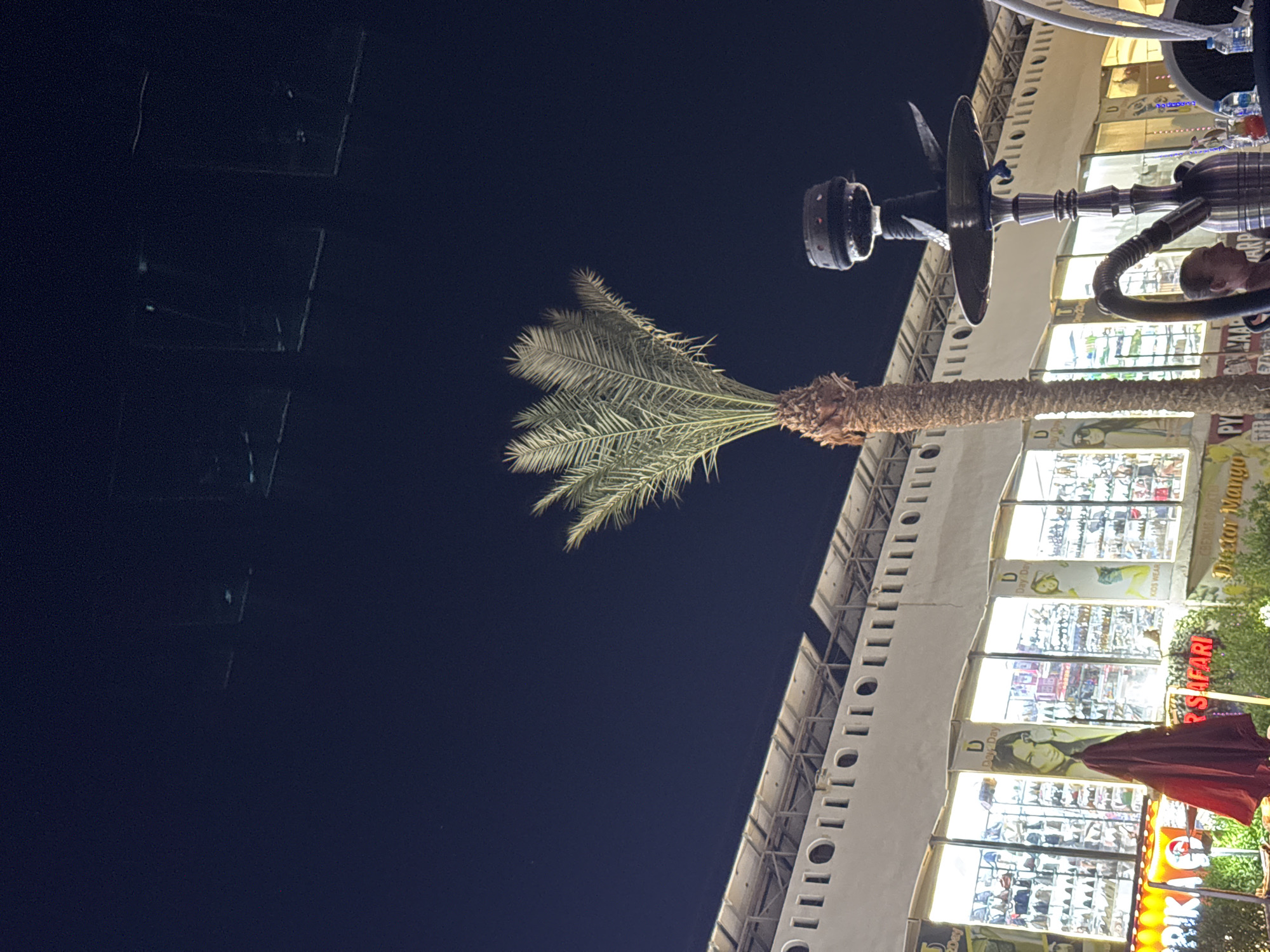 An exterior nighttime scene shows a low-angle view of a building with a row of shops and a palm tree in the foreground. The building is white with a decorative cornice and a row of windows displaying merchandise. Above the shops, solar panels are visible. To the right of the palm tree, a shisha pipe with a metal stem and a dark bowl is prominently featured. A person is visible in the lower right corner, facing left. The sky is dark blue and appears clear. Signs in the shop windows include "Day-Day" and "Safari". The overall atmosphere suggests a lively outdoor commercial area at night. The location is identified as Unknown, Egypt.