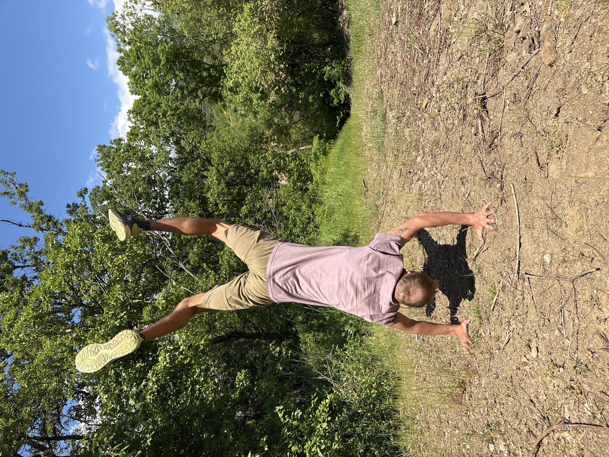A man is performing a handstand on a dirt path surrounded by trees and grass. The man is wearing a light purple t-shirt, khaki shorts, and green athletic shoes. The sky is visible through the trees and is a clear blue with a few white clouds. The location of the image is not in the city of Nizhnee Zaprudnoe, Ukraine.