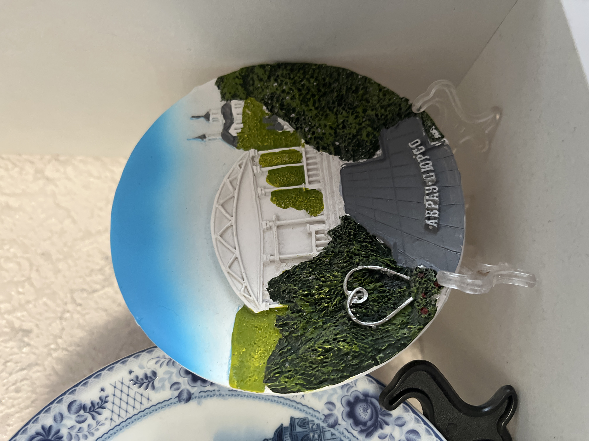 A circular, decorative plate depicting a scenic view of a building in Krasnyye Barrikady, Russia, is displayed on a white shelf. The plate features a white, ornate building with columns and a domed roof, surrounded by lush green trees and hills under a bright blue sky. The building appears to be a cultural or historical landmark. In the foreground, a paved pathway leads towards the building. A silver wire heart embellishment is placed on the left side of the plate. The plate is supported by a clear plastic stand. To the left of the plate, a portion of another decorative plate with a blue and white floral pattern is visible. A black plastic stand is also present on the shelf. The background is a textured white wall.