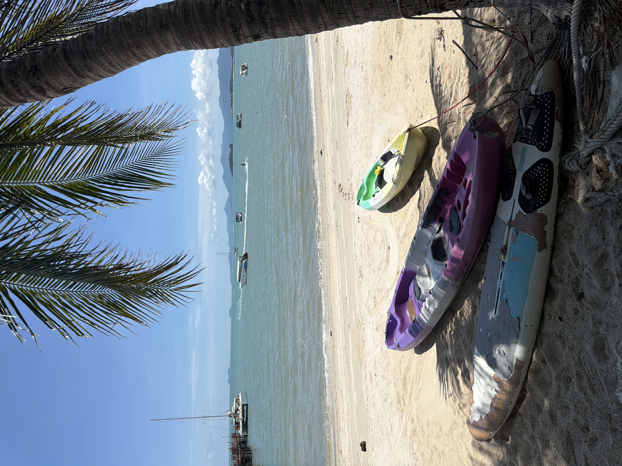 A beach scene in Baan Bang Rak, Thailand, depicts several watercraft on a sandy shore under a bright blue sky with scattered clouds. In the foreground, three watercraft are resting on the sand. The closest is a weathered surfboard with peeling paint, showing shades of brown, white, and blue, with a pattern of black dots near its nose. Behind it is a purple and grey kayak, and further back, a green and yellow kayak. The ocean stretches out behind the beach, with gentle waves lapping the shore. Several boats are visible on the water, including a large white catamaran docked at a wooden pier to the left, and smaller motorboats further out. Palm tree fronds hang from the right side of the frame, casting shadows on the sand. The overall impression is a tranquil tropical setting.