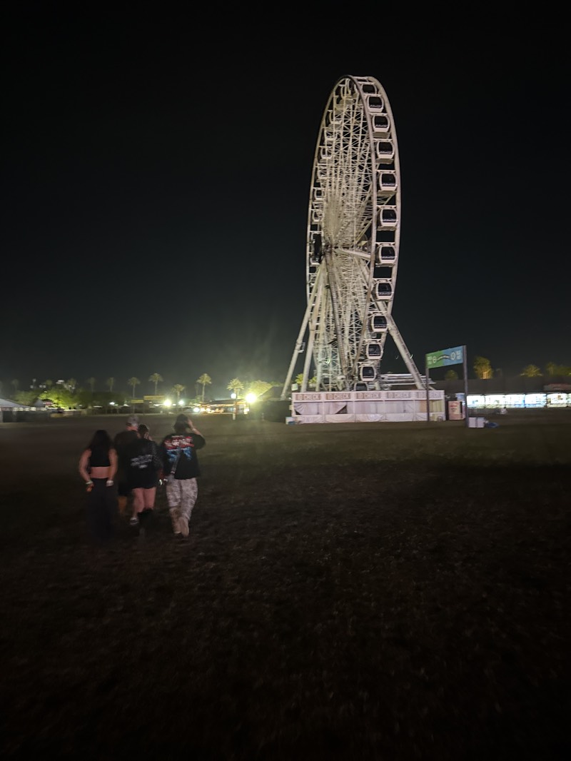 The image depicts a night scene at what appears to be a festival or large event in Indio, United States. A Ferris wheel is the prominent structure, towering over the area, illuminated against the dark sky. Several people are walking across a field, away from the viewer, in the direction of the Ferris wheel. The ground appears to be grassy or possibly muddy, with light poles and structures visible in the background, suggesting other festival amenities. The overall environment seems to be dimly lit, consistent with a nighttime outdoor event.