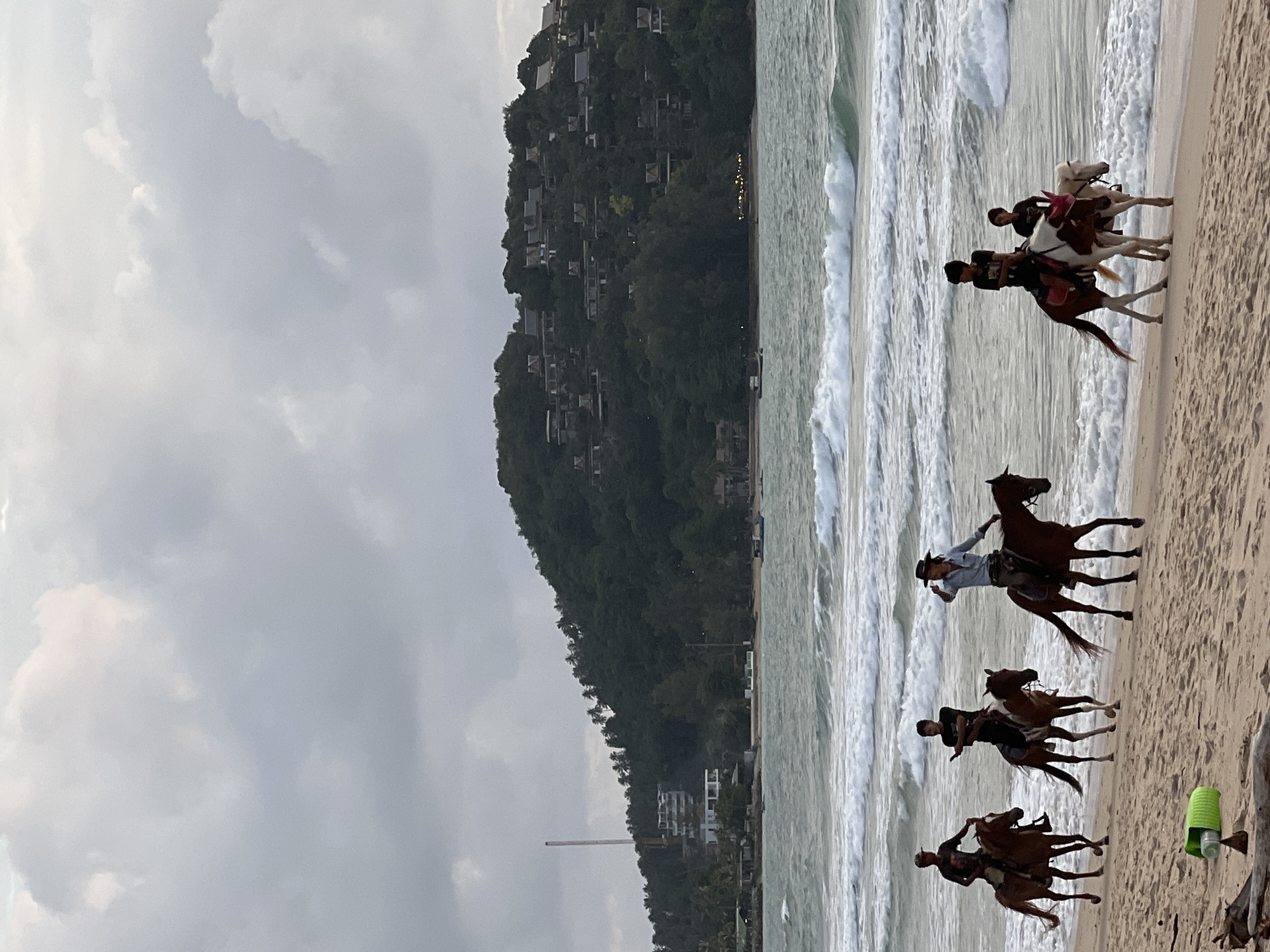 The image depicts a beach in Choeng Thale, Thailand, under an overcast sky. Five people are riding horses along the shoreline, with the ocean and waves in the background. The sand is a light brown color, and there are some pieces of driftwood and a green plastic bottle on the beach.
In the foreground, there are three people riding horses. The first person on the left is riding a brown horse. The second person is riding a brown and white horse, and appears to be holding a phone. The third person is wearing a cowboy hat and riding a brown horse.
Further down the beach, closer to the water, two more people are riding horses. One is on a brown and white horse with a pink bridle, and the other is on a white horse.
The ocean is a greenish-blue color with white waves crashing on the shore. In the distance, a lush green hillside is covered in trees and dotted with buildings, suggesting a populated area or resort. The sky is a muted grey, indicating cloudy weather.