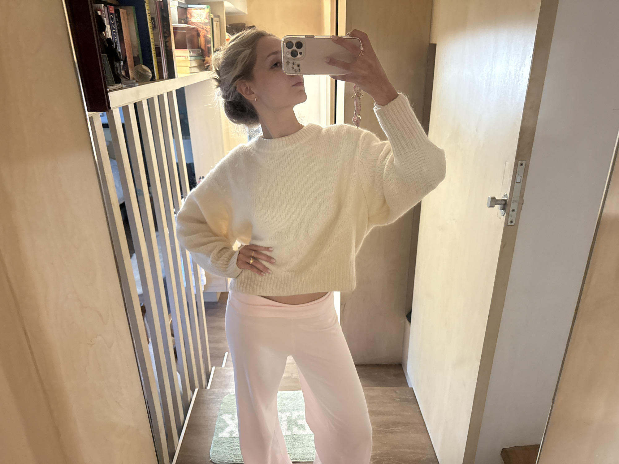 A young woman with blonde hair tied up in a bun takes a mirror selfie indoors. She is wearing a cream-colored oversized sweater and light pink wide-leg pants. She is standing in what appears to be a room with wooden walls and a staircase with light-colored railings. There are bookshelves in the background and a door with a silver handle on the right side of the frame. The floor is made of wooden planks, and a green rug is visible under the staircase. The lighting in the room is warm and natural, coming from a source not visible in the frame. The overall scene is casual and domestic, possibly a bedroom or dressing area. There is no indication of the specific location within Ciudad de México, Mexico.