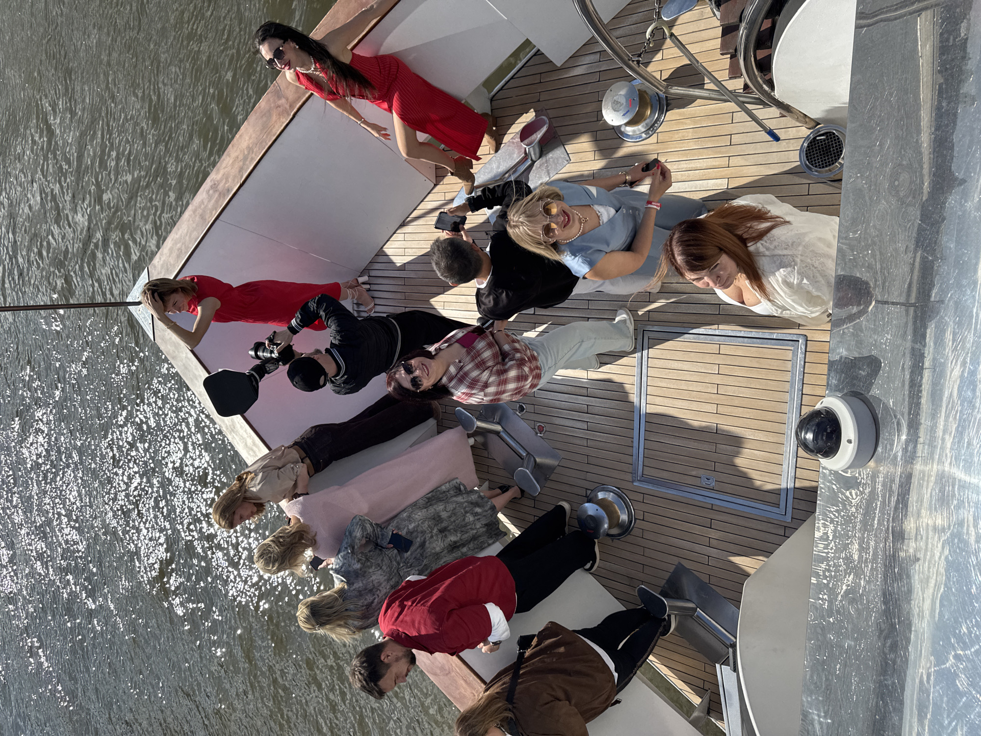 On a yacht in Moscow, Russia, a group of people are gathered. Two women in red dresses are reclining on a white bench near the edge of the yacht, close to the water. A photographer is positioned nearby, capturing their image. Adjacent to them, a man in a black jacket and a woman in a red and white plaid shirt are sitting. The others are also relaxing. The deck of the yacht is made of wood. In the background, the city of Moscow is visible.