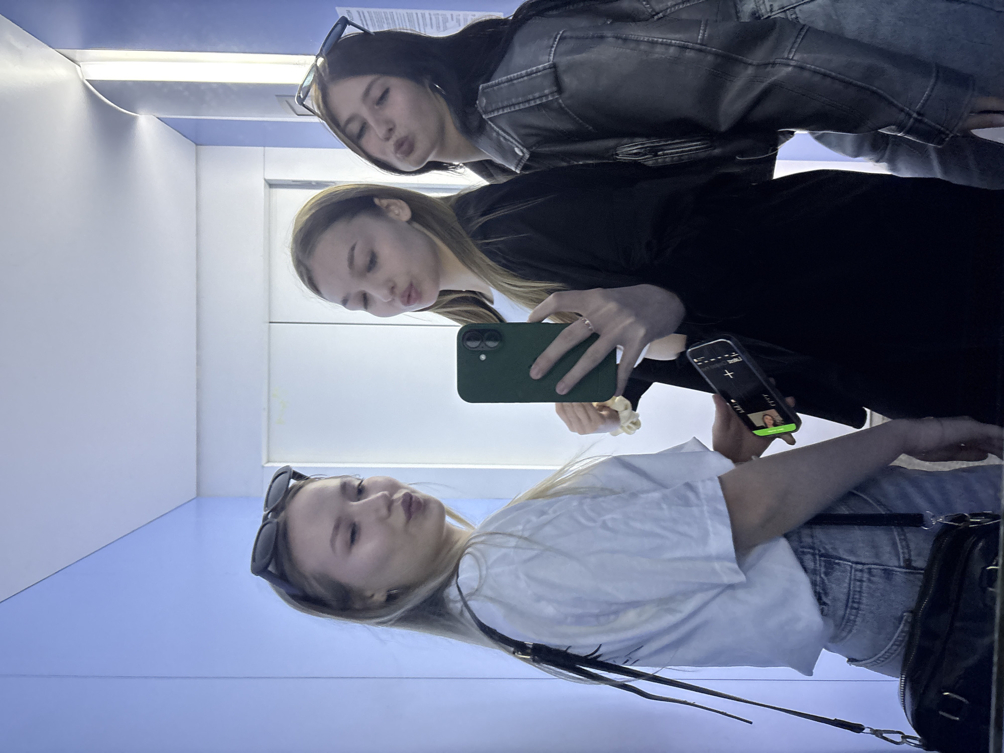 Three young women are taking a mirror selfie in a narrow, modern hallway with blue walls. The woman on the left, with blonde hair and sunglasses pushed up on her head, wears a white t-shirt and denim shorts, and carries a black purse. The woman in the middle, with brown hair, wears a black top and holds a green smartphone, taking the photo. The woman on the right, with dark hair and sunglasses on her head, wears a denim jacket over a black top. They are all making duck faces for the camera. The location is not specified beyond "in the city of Chaikovsky, Russia."