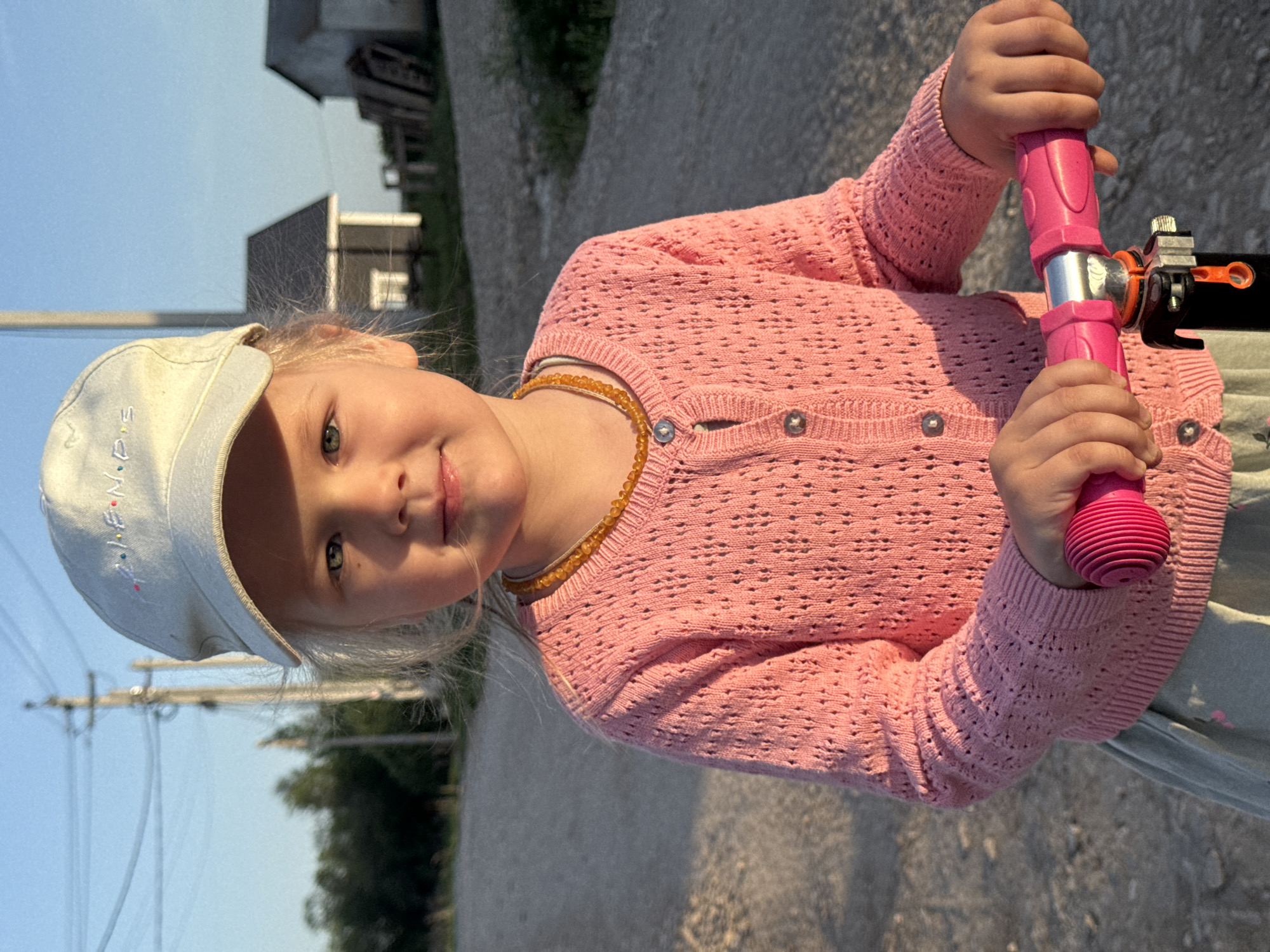 A young girl in Ферма, Russia, wearing a white baseball cap, a pink cardigan, and a beaded necklace, stands on a gravel road holding the pink handlebars of a scooter. In the background, there is a building and some greenery. The sky is blue.