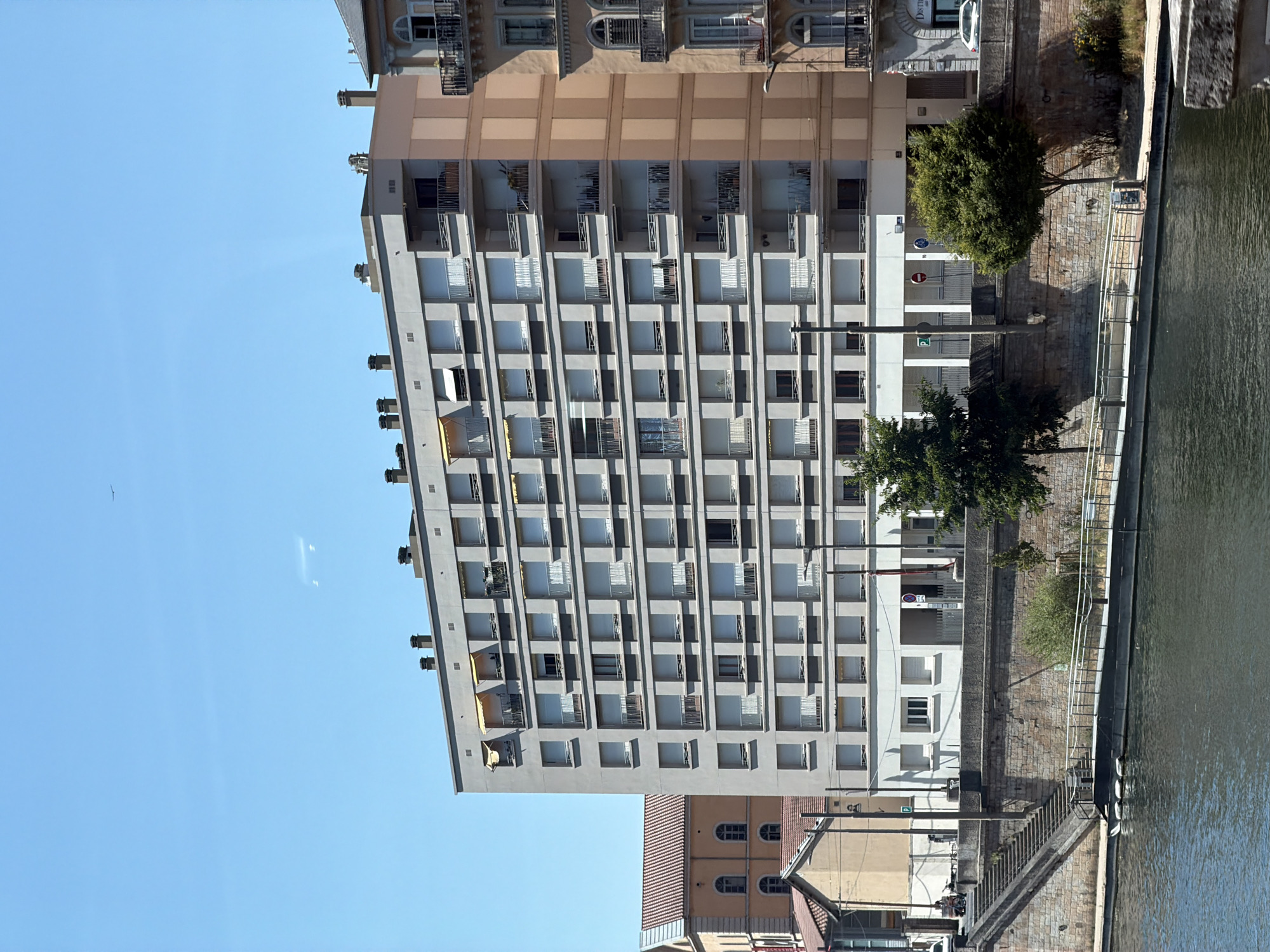 The image captures a bright, sunny day in Lyon, France, looking across a body of water towards a residential building complex. The primary structure is a multi-story apartment building with a light gray facade and numerous balconies, each equipped with horizontal railings and roller shutters. The building's windows are also largely covered by these shutters, giving it a uniform appearance. On the far right, another building with a light brown facade features ornate balconies with wrought-iron railings and arched windows. Adjacent to these, a building with a terracotta-tiled roof and pale orange walls has several windows, some of which appear to be arched.
The foreground is dominated by the dark, rippling water, likely a river, reflecting the clear blue sky. Along the waterfront is a stone embankment topped by a walkway with metal railings. Stairs lead up from the water's edge to this walkway. A few trees are visible, including a large, leafy green tree situated near the apartment building and another smaller tree to its left. The environment appears to be urban, with a clear sky and strong sunlight casting shadows. There are no prominent individuals or specific actions being depicted, emphasizing the architectural and environmental aspects of the scene. A single bird is visible as a small dot in the upper sky.