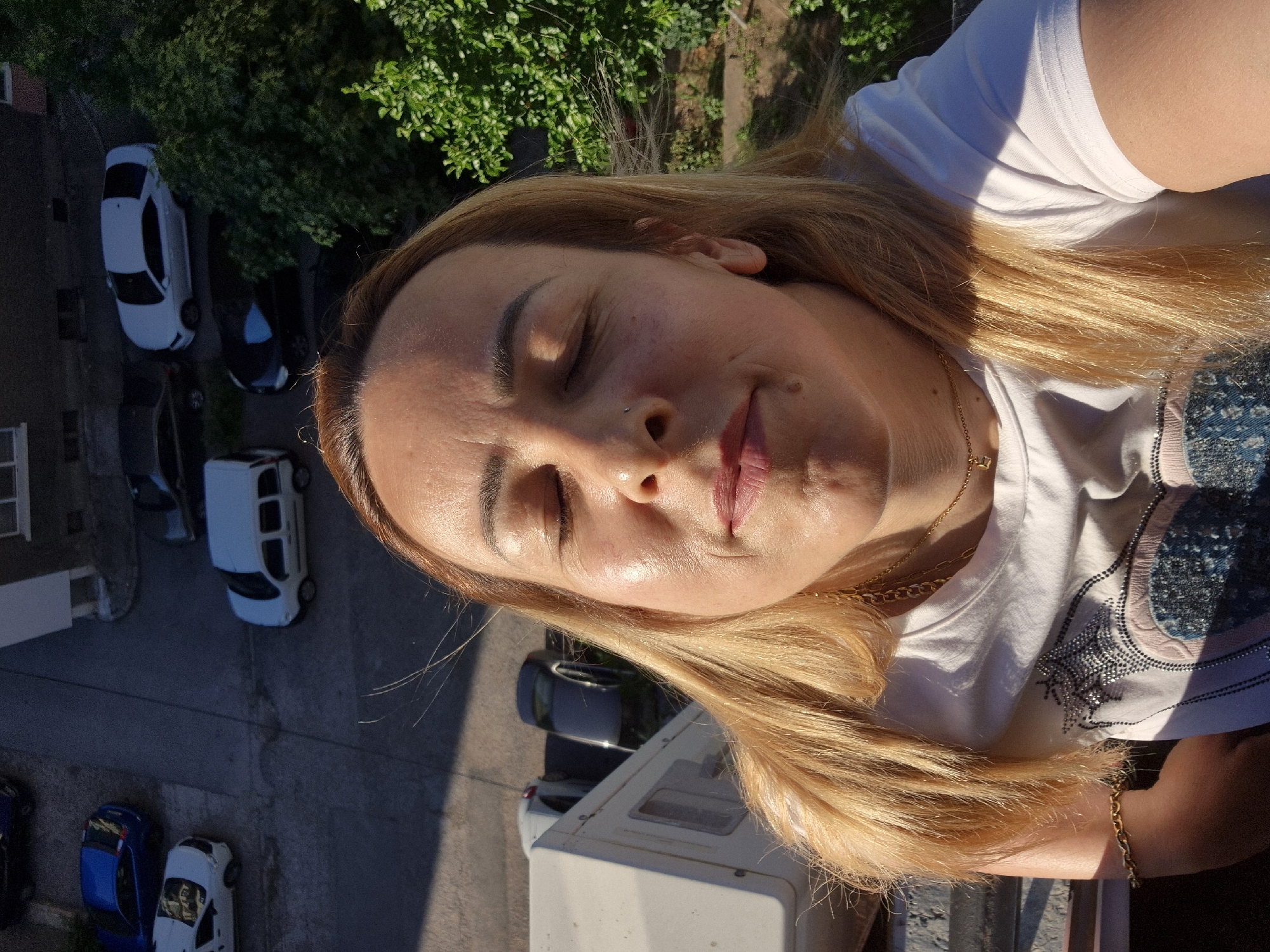 A woman with blonde hair and closed eyes is shown in a close-up selfie from a high angle. She is wearing a white t-shirt with a graphic print and gold jewelry, including a necklace and bracelet. Sunlight illuminates her face, highlighting her skin. In the background, a street with several parked cars and a tree can be seen. The perspective suggests the photo was taken from a balcony or upper floor of a building, overlooking a residential area.