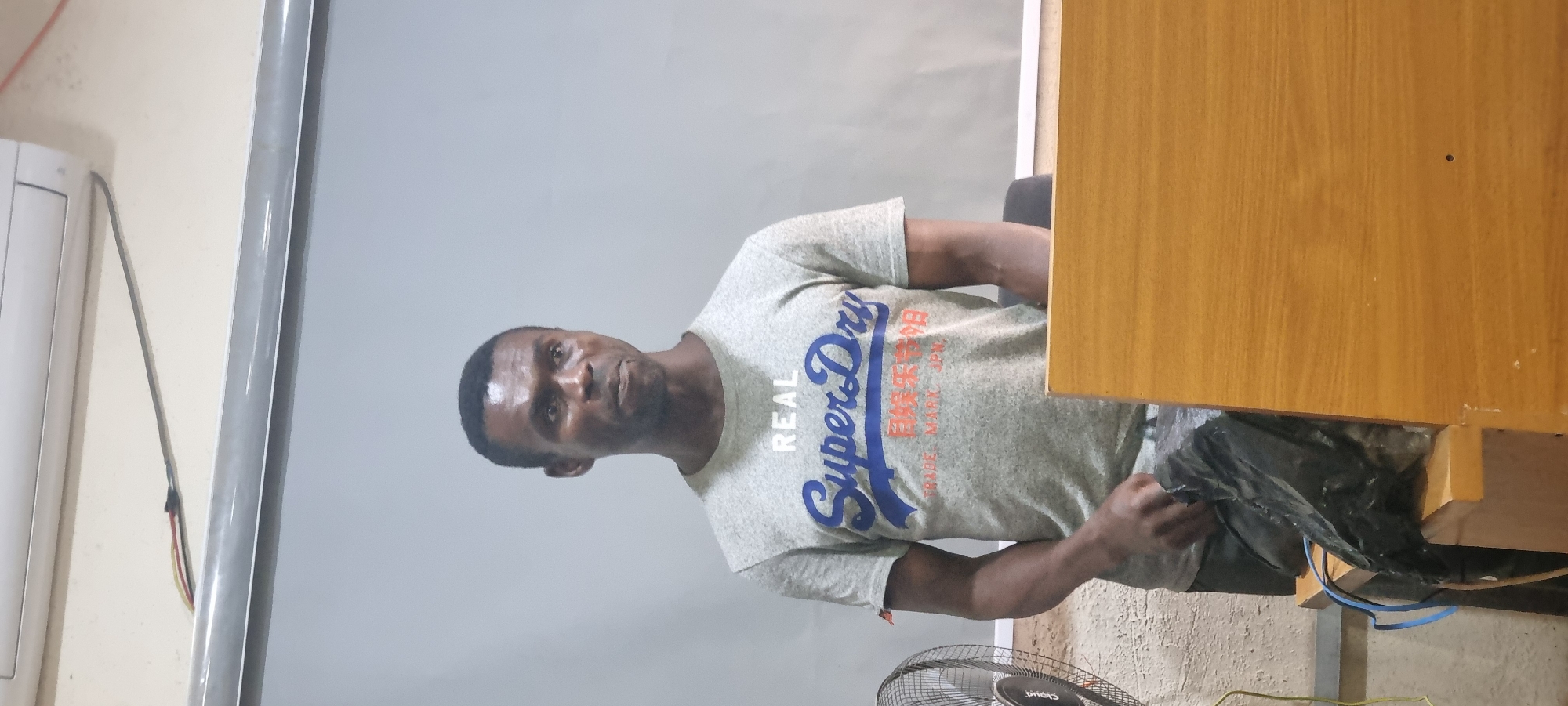 A Black male stands in front of a light gray projection screen, wearing a light gray t-shirt with blue and red Japanese characters and "Super Dry" printed on it. He is holding a black plastic bag in his left hand. A wooden desk is in front of him, and a black fan is to his left. An air conditioner is mounted on the wall above him. The scene appears to be indoors, possibly in a classroom or meeting room.