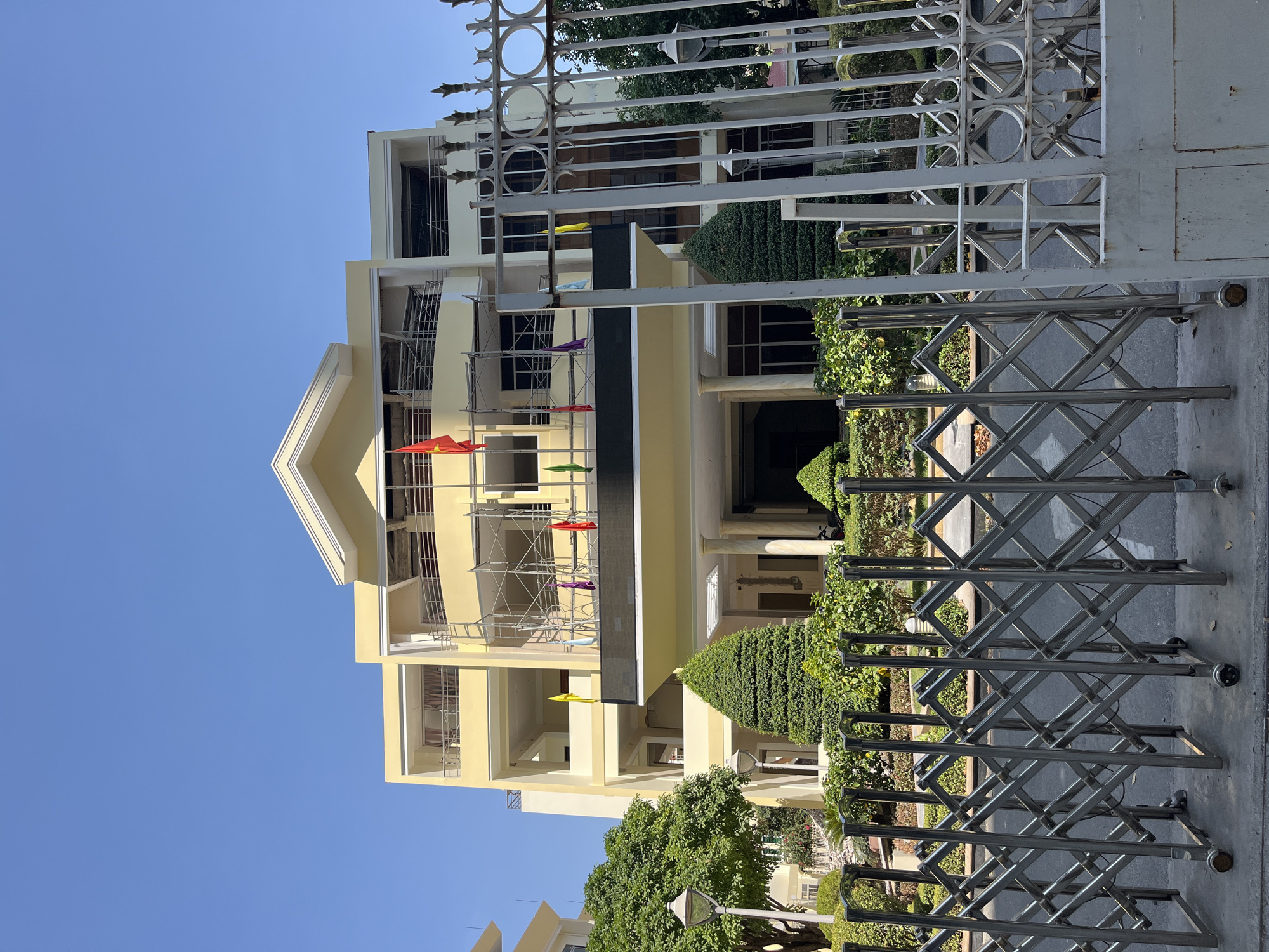 The image shows a light yellow building in Nha Trang Ward, Vietnam, viewed through two layers of metal gates. The building has multiple floors, a triangular roofline, and scaffolding with colorful flags attached. Landscaped greenery is visible in front of the building. The sky is clear and blue.