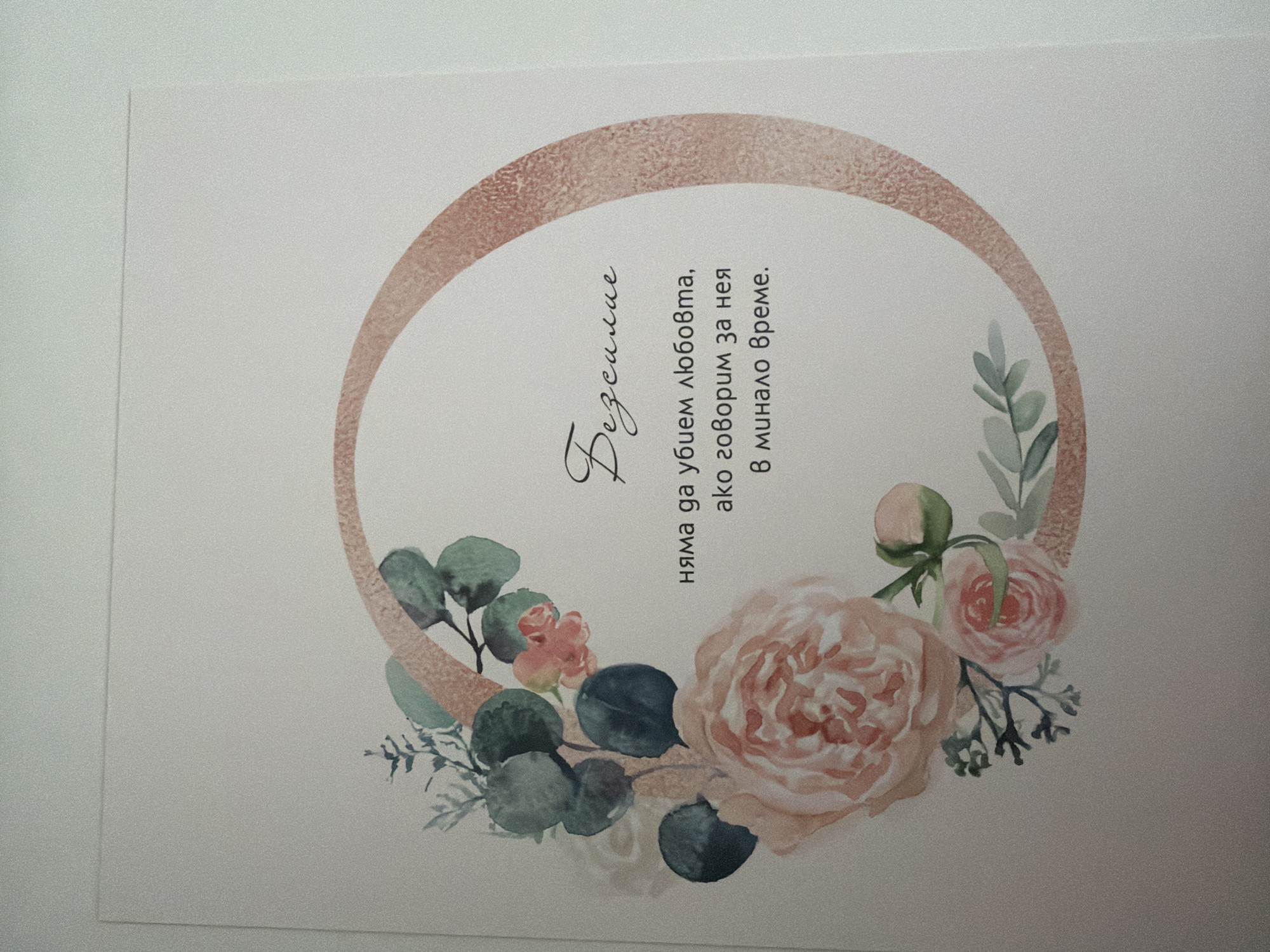 The image shows a close-up, top-down view of a card with floral illustrations and text. A large, pale pink rose is prominent on the left, with smaller pink roses and green leaves arranged around it. A rose-gold colored, textured circle frames text in the center of the card. The text, written in a cursive font, reads: "Безсилие няма да убием любовта, ако говорим за нея в минало време." This translates from Bulgarian to English as: "Helplessness we will not kill love, if we speak of it in the past tense." The card is placed on a plain, off-white surface. There is no discernible location in the city of Pazardzhik, Bulgaria depicted in the image. The image is a still photograph with no visible action or interaction.