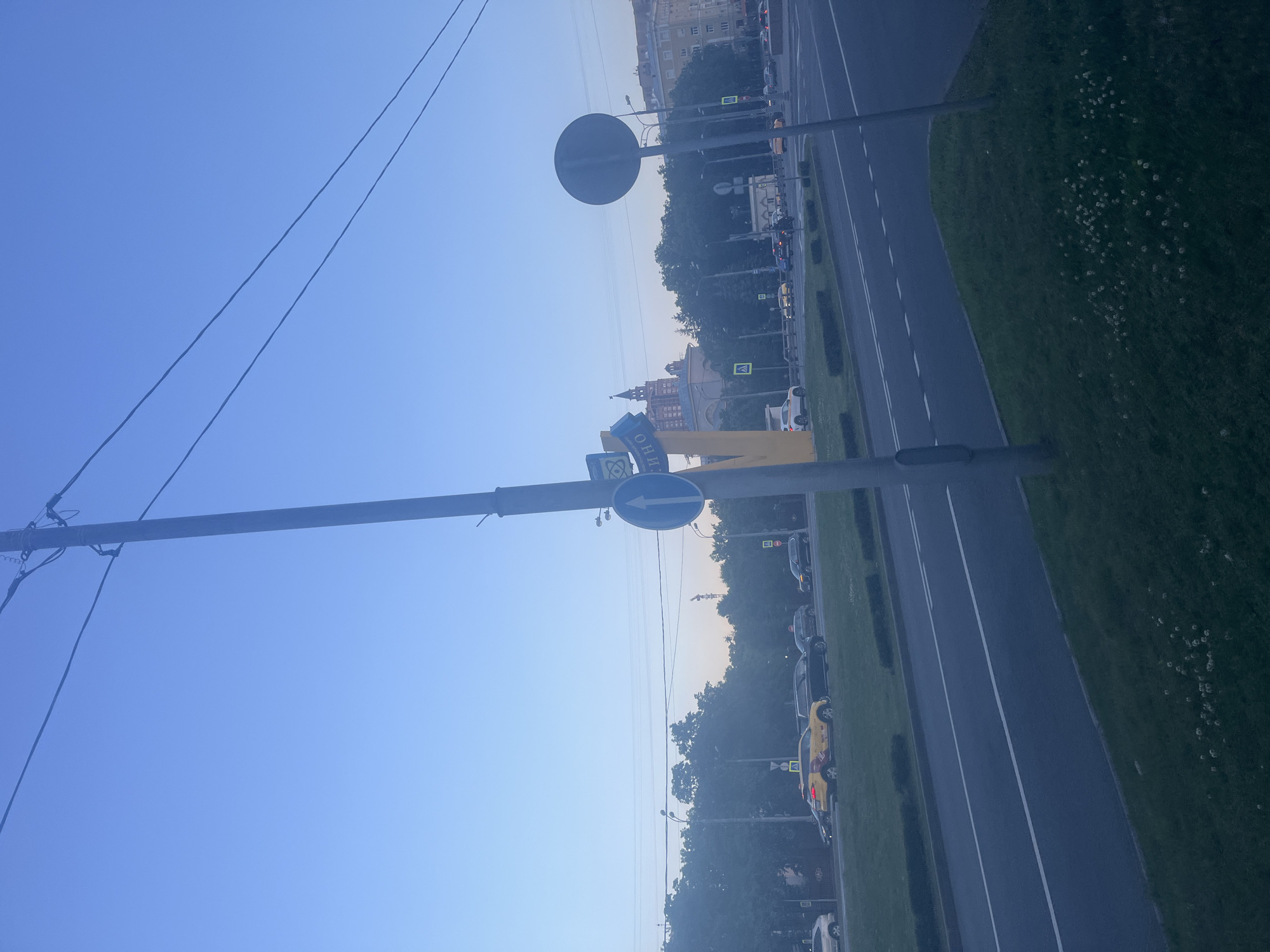 This is a medium-angle, eye-level shot of a street scene in Khimki, Russia, captured in the early morning or late evening due to the soft, diffused light. The sky is a clear, pale blue, transitioning to a lighter hue near the horizon. A tall, grey utility pole dominates the foreground, with several dark electrical wires extending diagonally across the upper portion of the frame. To the right of the pole, a circular, dark grey traffic sign is visible. On the left side of the pole, a blue circular traffic sign with a white upward-pointing arrow is attached.
Further down the street, a yellow taxi is the most prominent vehicle, positioned in the middle lane of a multi-lane road. Other vehicles, including black cars and what appears to be a white car, are also visible on the road. A light-colored building with a distinct tower, possibly a cultural or administrative building, is visible in the background, partially obscured by trees. Several streetlights and traffic signs, including yellow pedestrian crossing signs, are scattered along the roadside. The scene includes a grassy median strip with sparse white flowers and a paved roadway. The overall impression is a quiet urban environment at twilight.